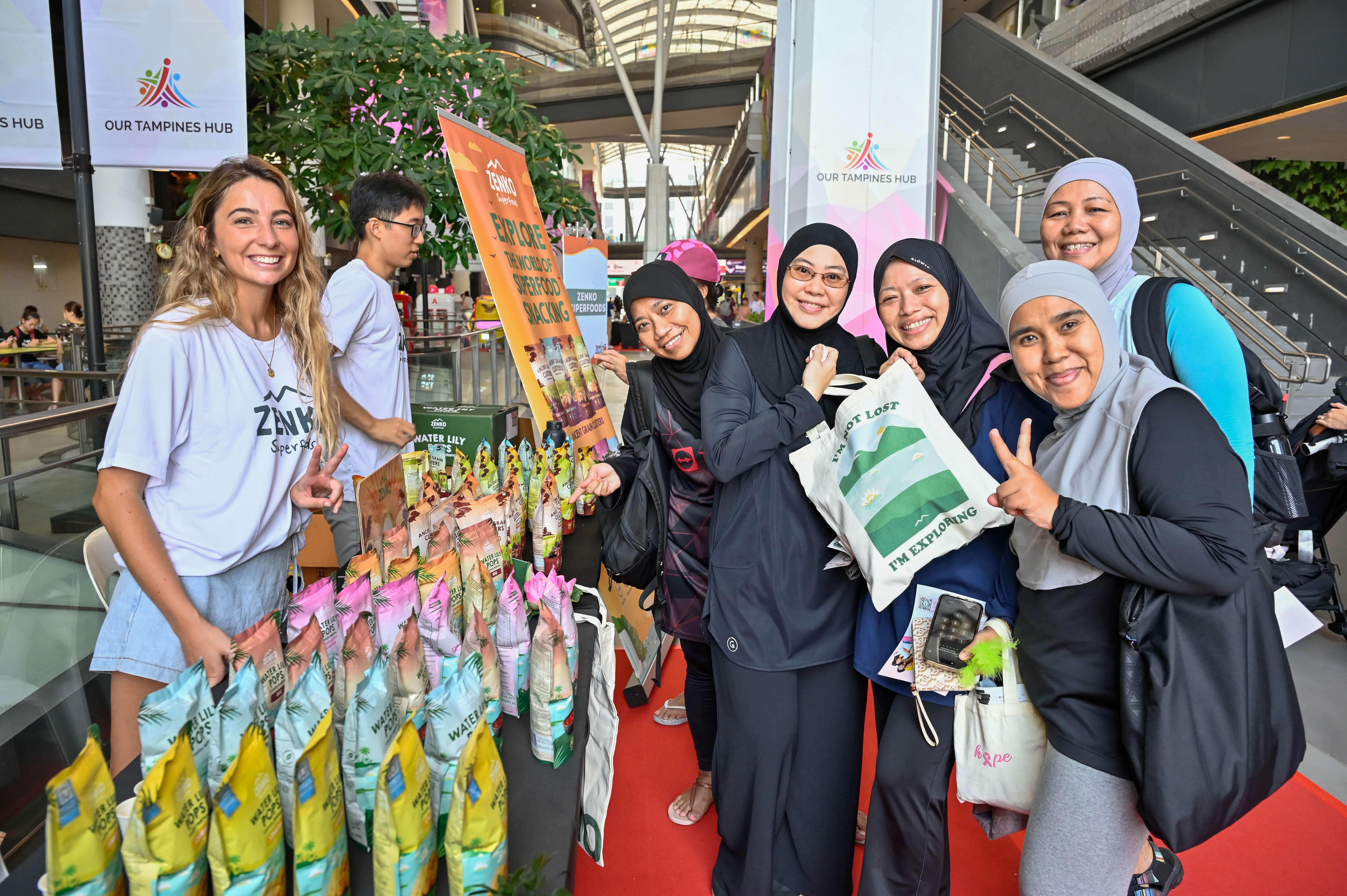 A group of people stands near a display of snacks at a booth in a shopping center with banners and stairs in the background.