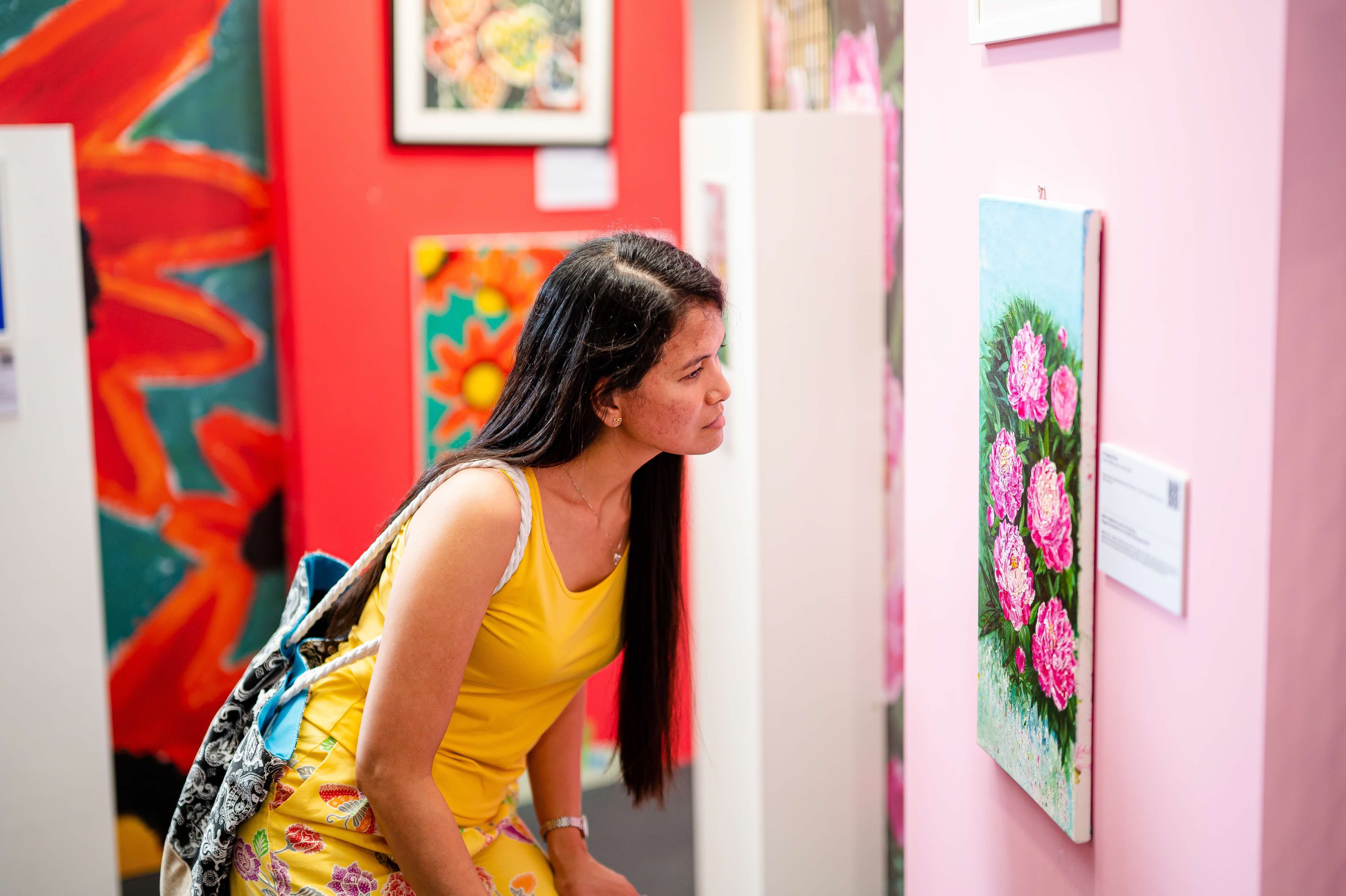 A woman in a yellow dress looks closely at a floral painting in an art gallery with colourful walls and exhibits.