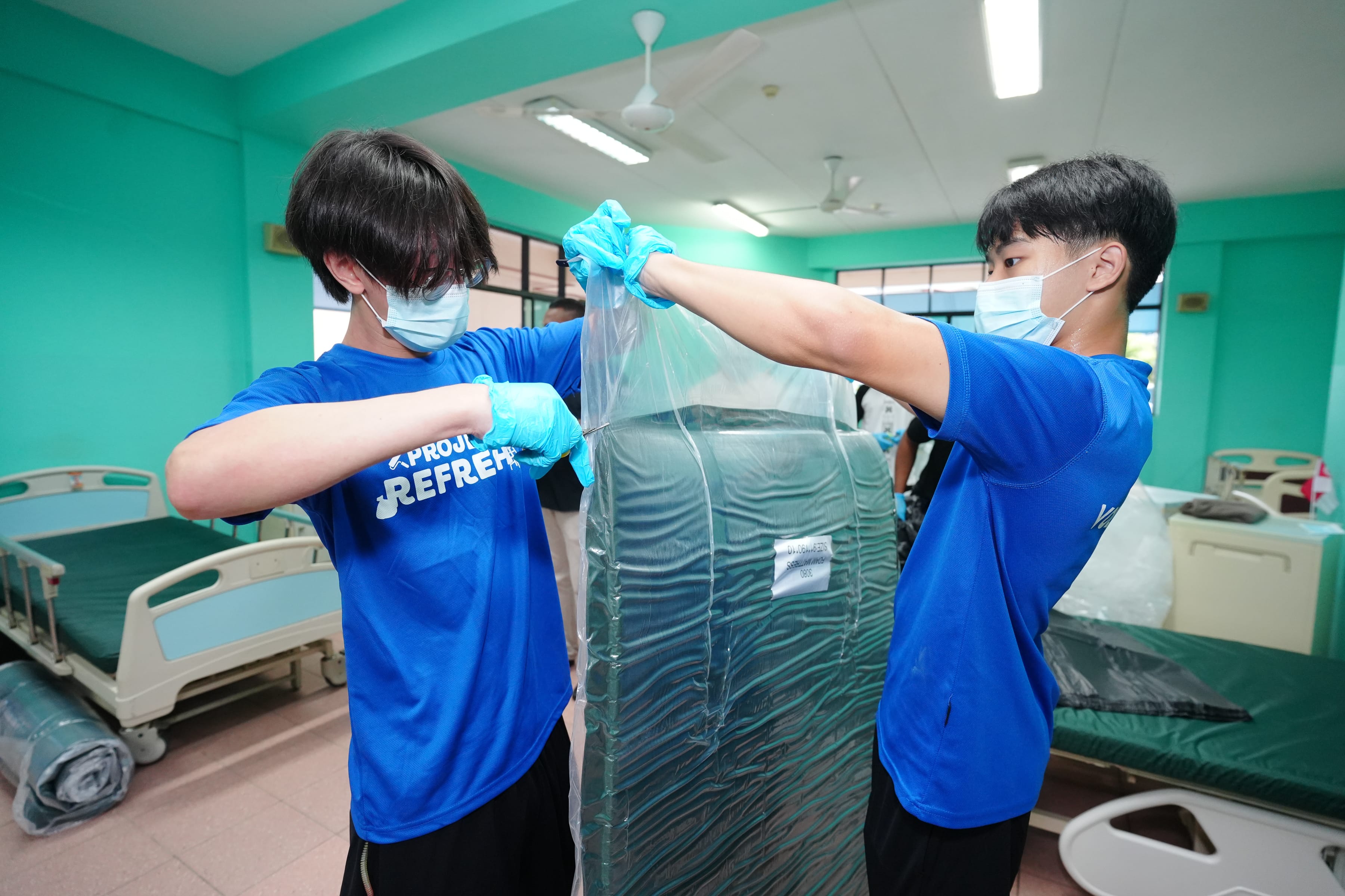 Two people in blue shirts and gloves are handling plastic-wrapped mattresses in a room with hospital beds.