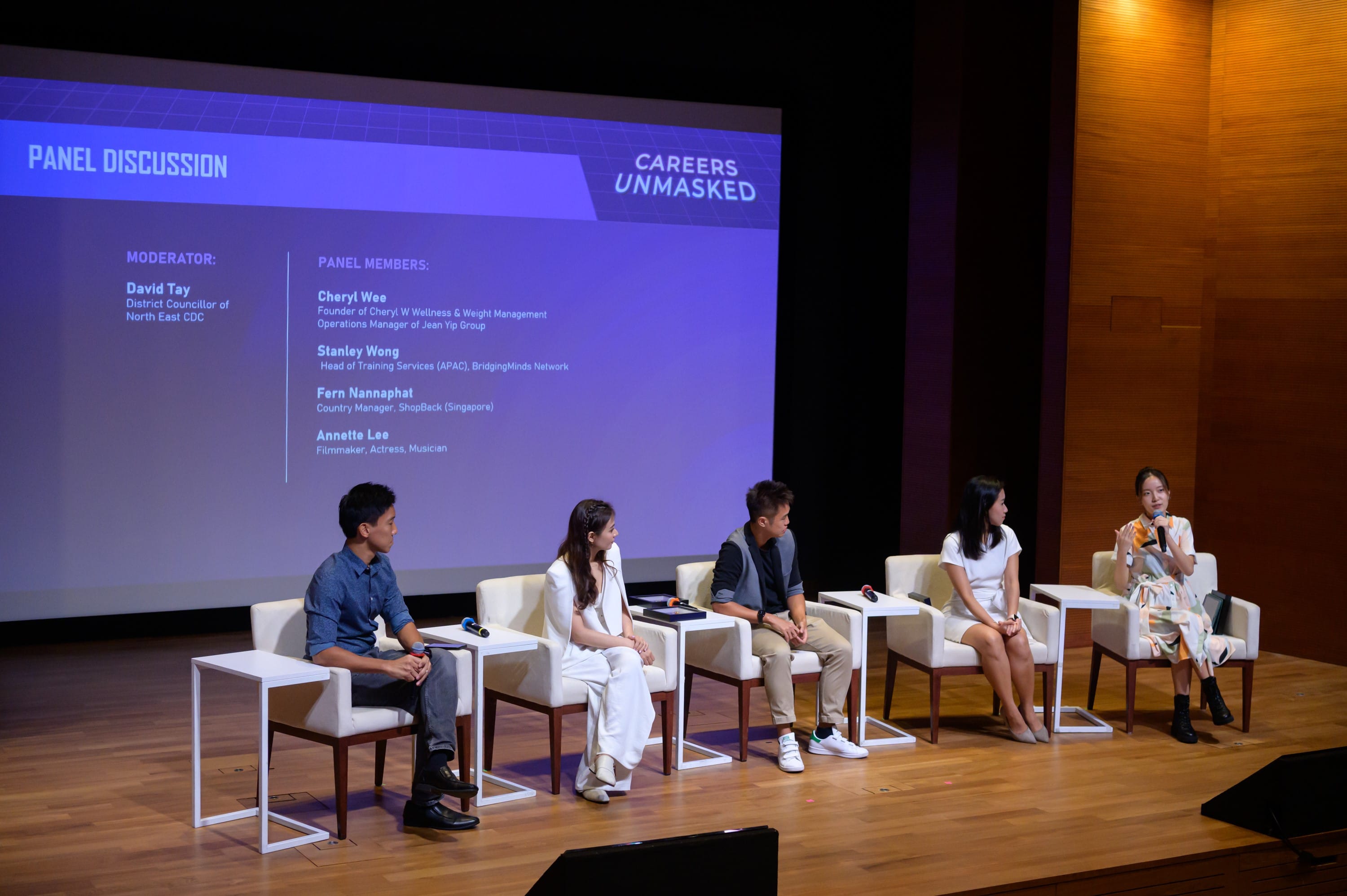 Five individuals sit on stage for a careers panel discussion, with a projected screen listing their names and titles.