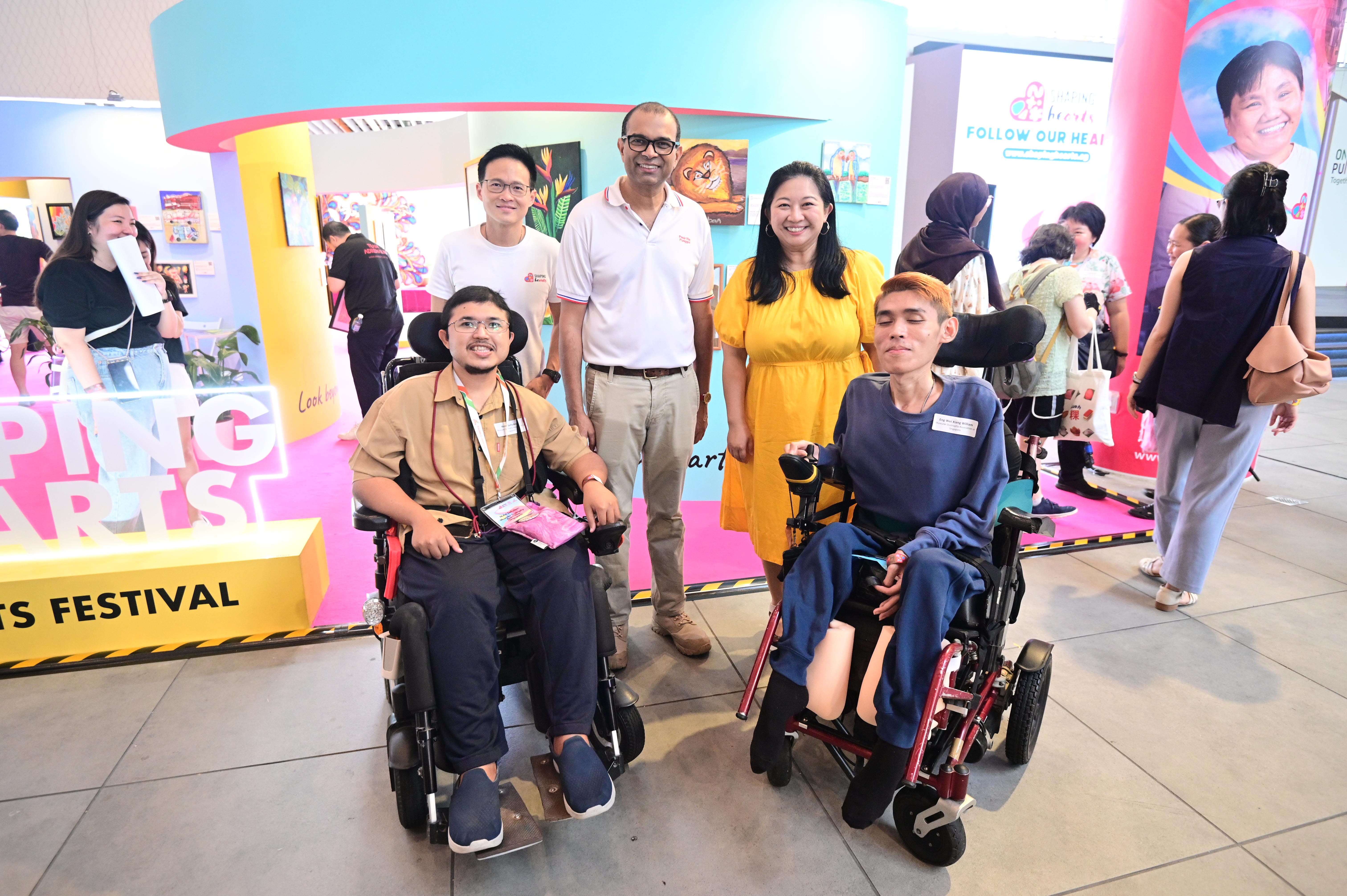 Group photo at an inclusive arts festival featuring participants and guests, including two people using wheelchairs, gathered in front of a colourful exhibition booth.