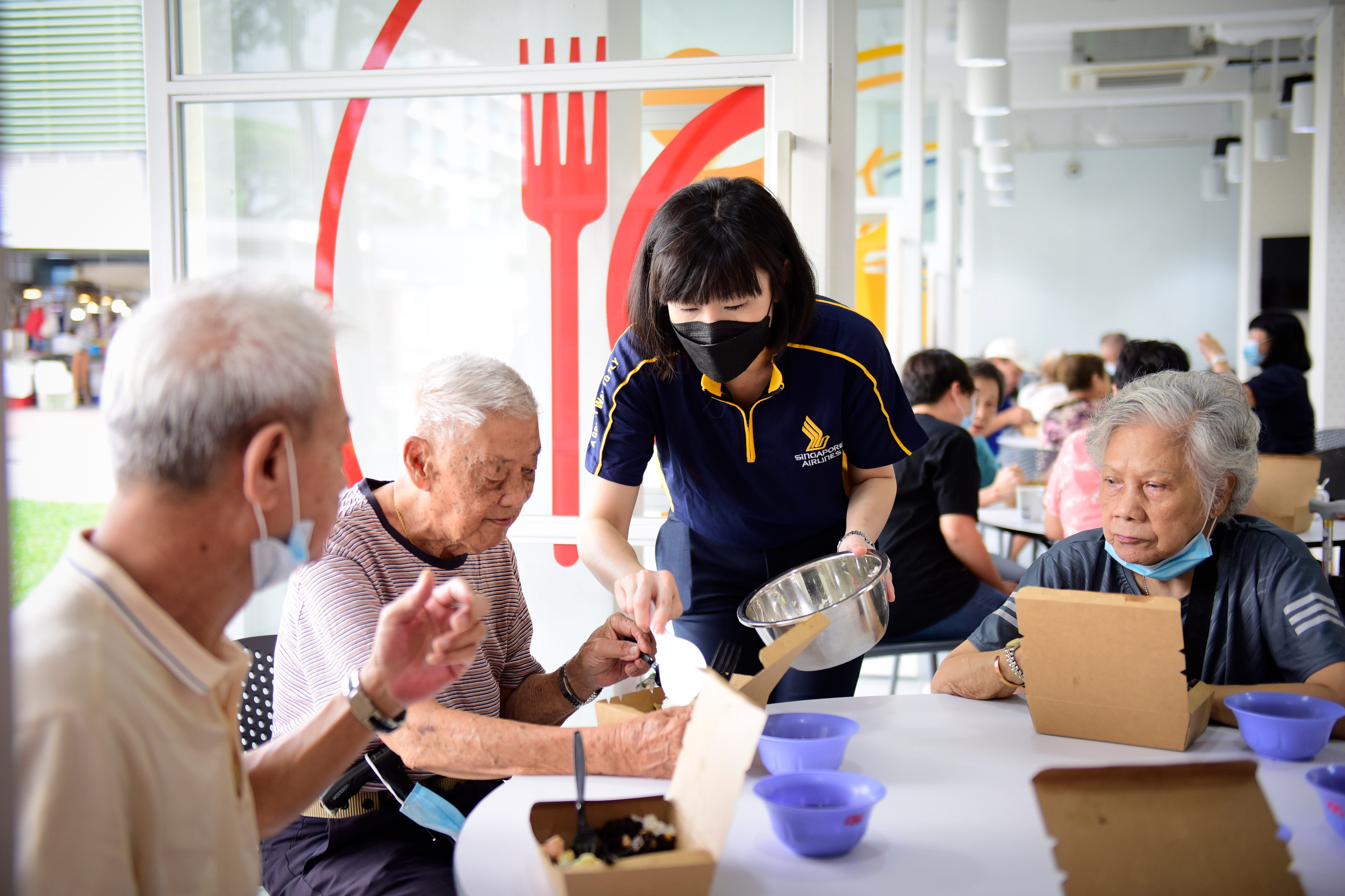 A worker assists elderly people with their meals at a community center, which features a large red fork design on the window.