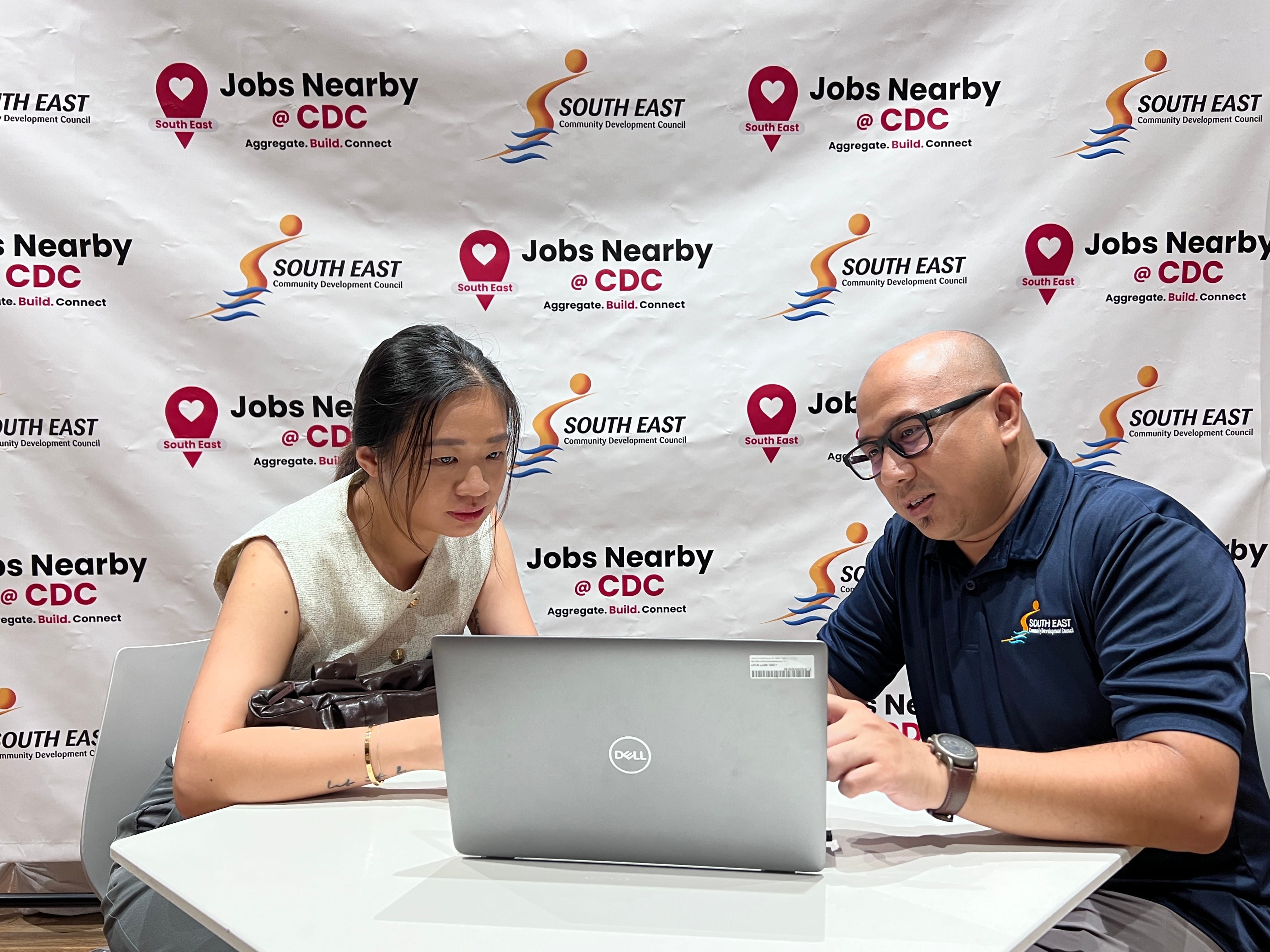 Two people sit at a table with a laptop in front of a backdrop featuring South East CDC and Jobs Nearby logos.
