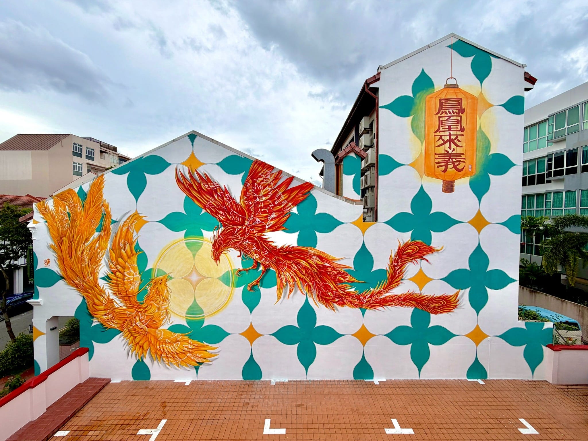 Large wall mural on the side of a building featuring two vivid phoenixes circling a glowing motif, with a hanging lantern bearing Chinese characters against a patterned turquoise-and-white background.