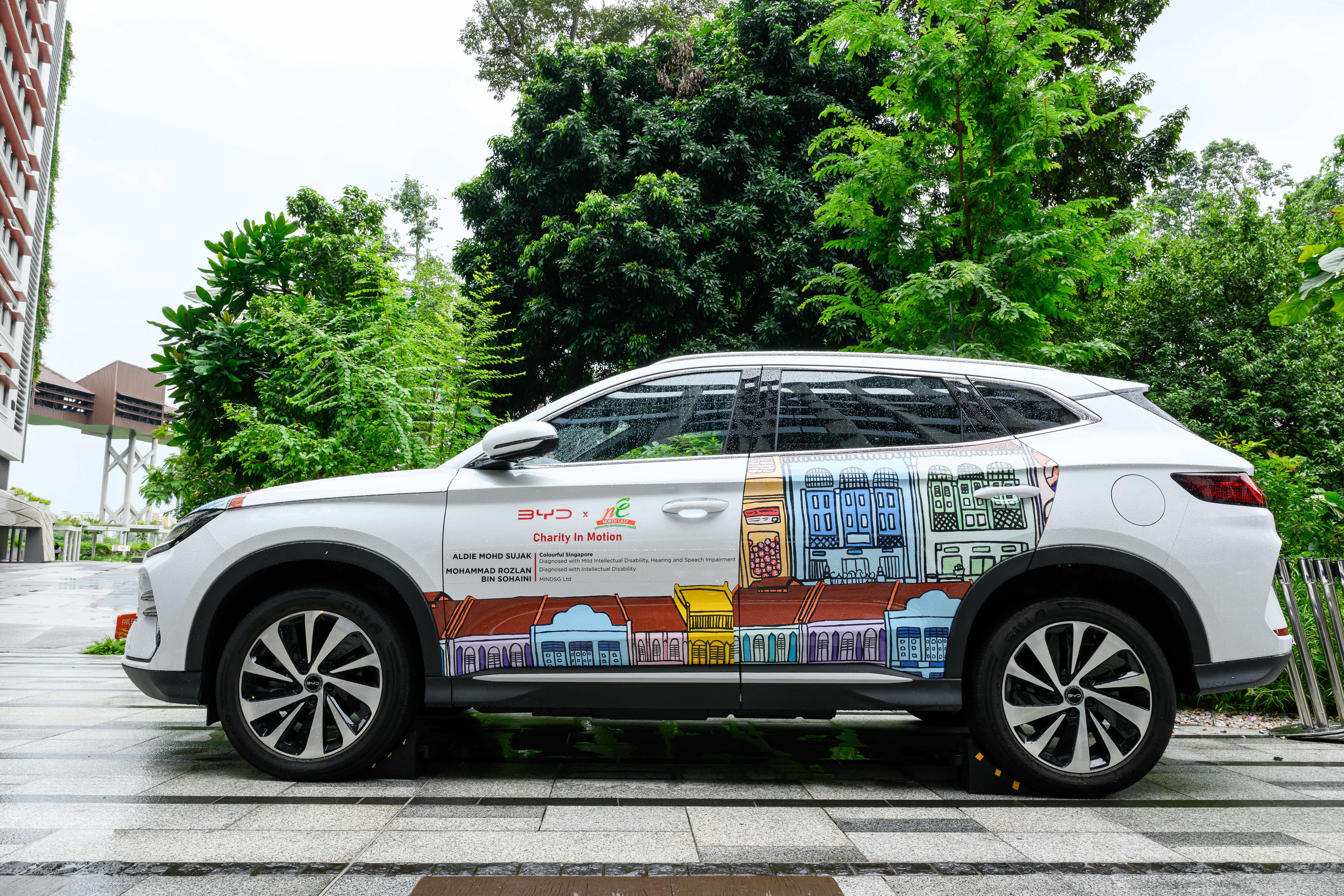 A white SUV parked outdoors is decorated along its side with colorful illustrated shophouses and text promoting a charity initiative called “Charity in Motion,” set against a backdrop of lush greenery and buildings.