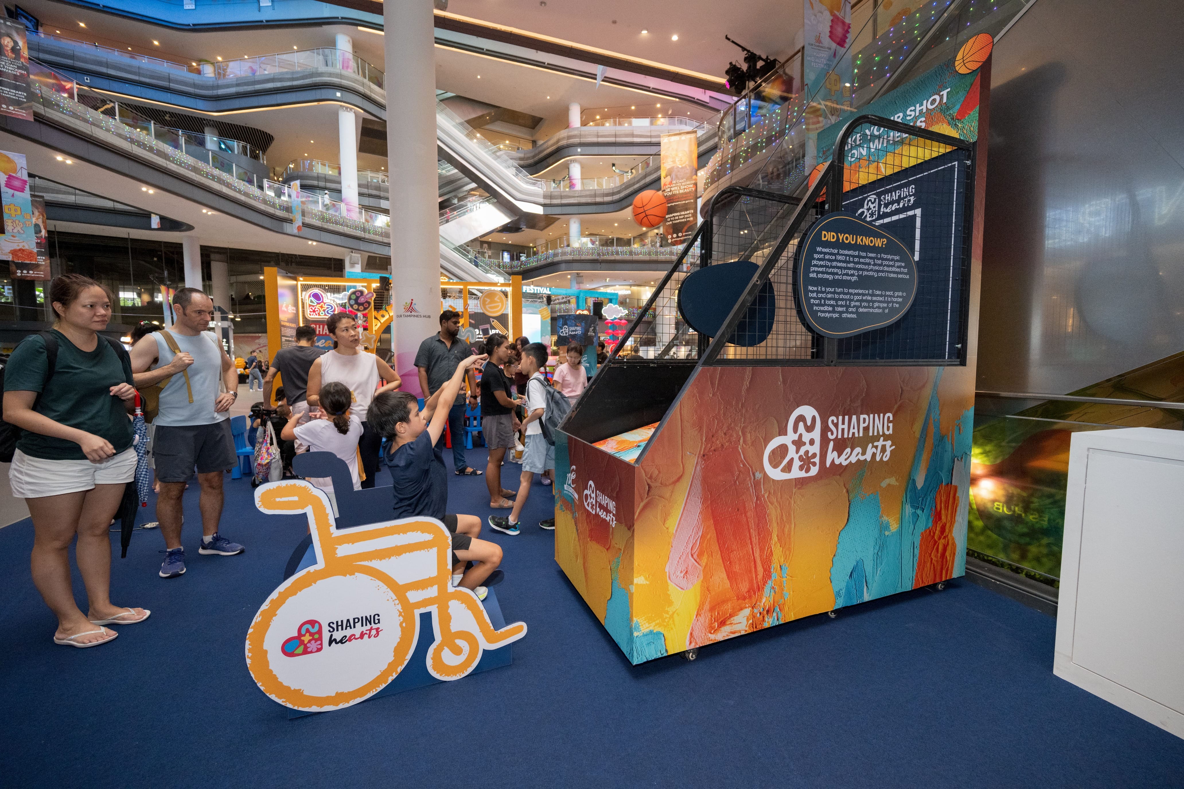 Boy sitting in a wheelchair-simulated chair, shooting a basketball into the hoop while his family watches from behind.