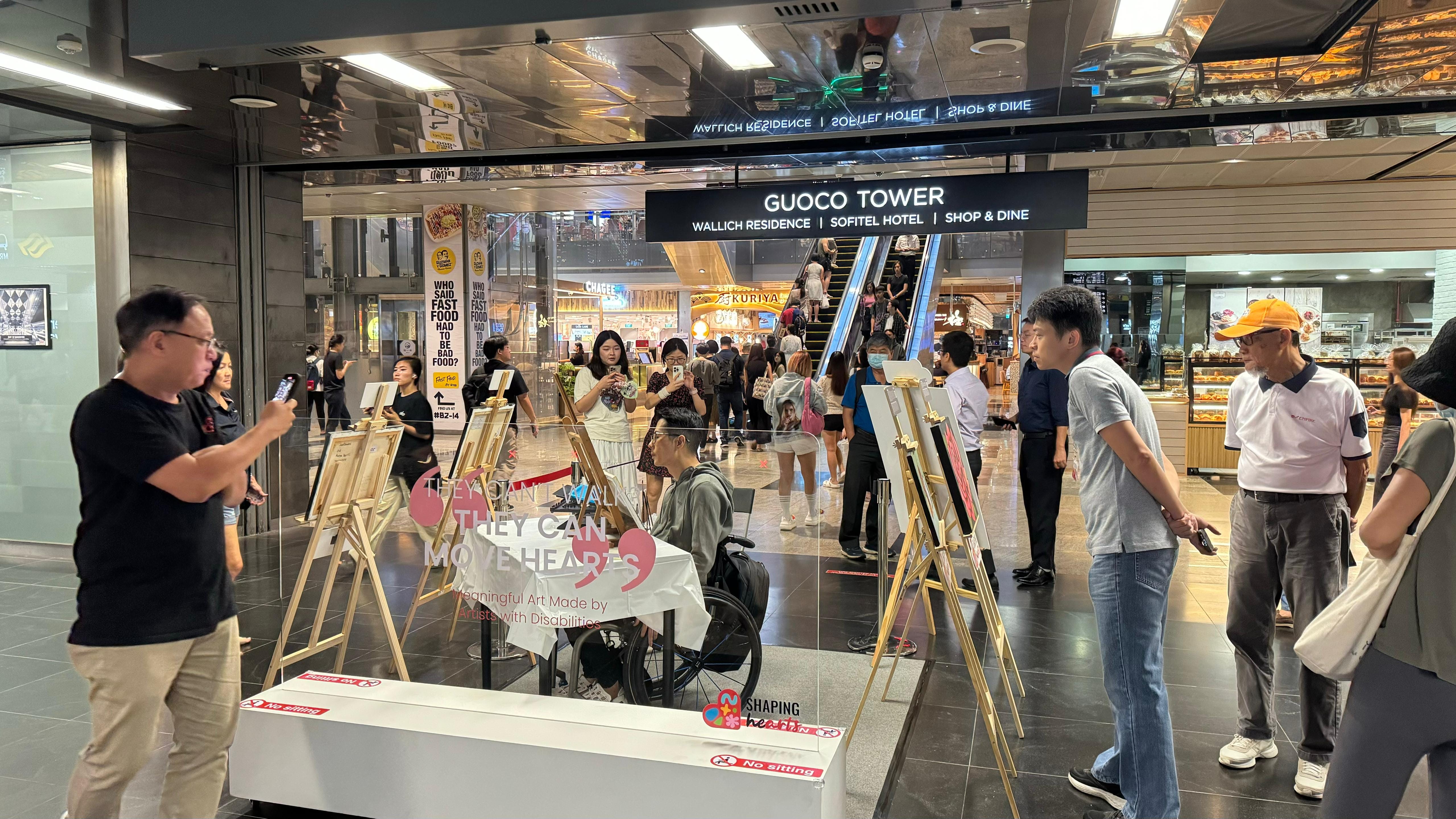 People are observing paintings displayed on easels in the corridor of a shopping mall.