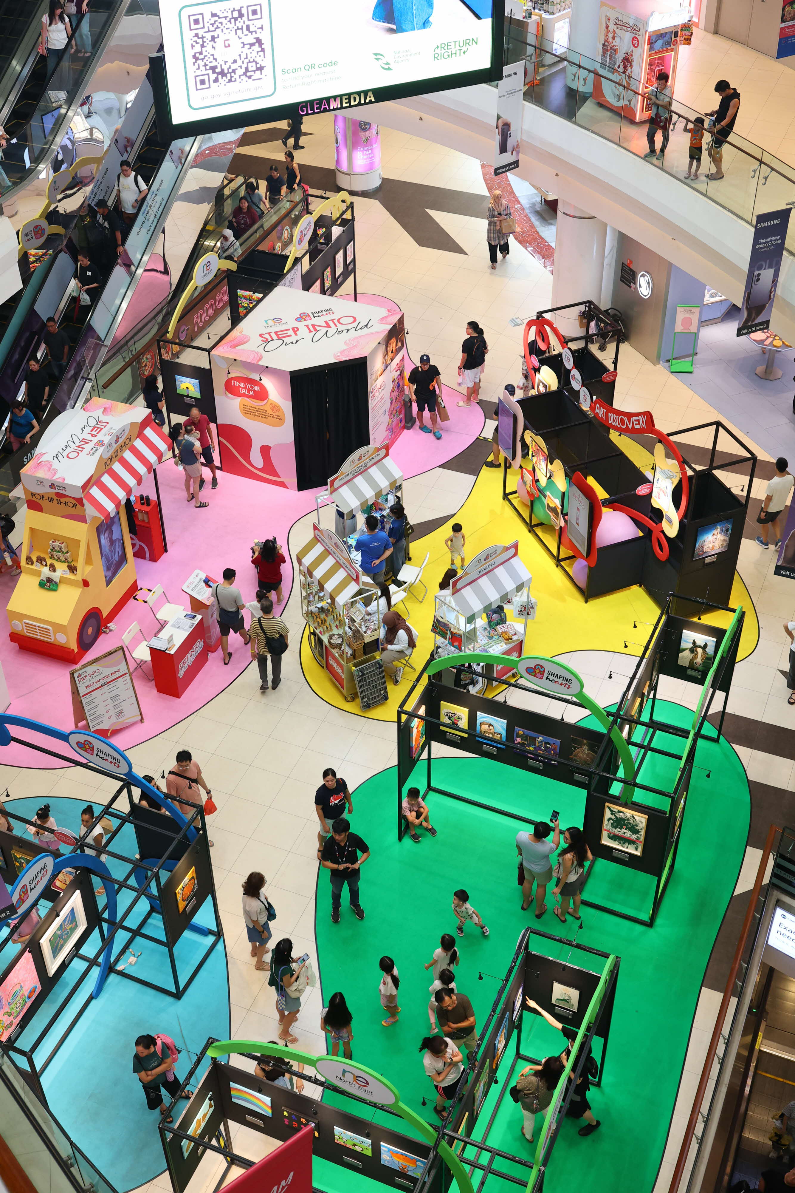 An overhead view of the Shaping Hearts exhibition at Tampines Mall, featuring art displays, activity booths and residents exploring the event.