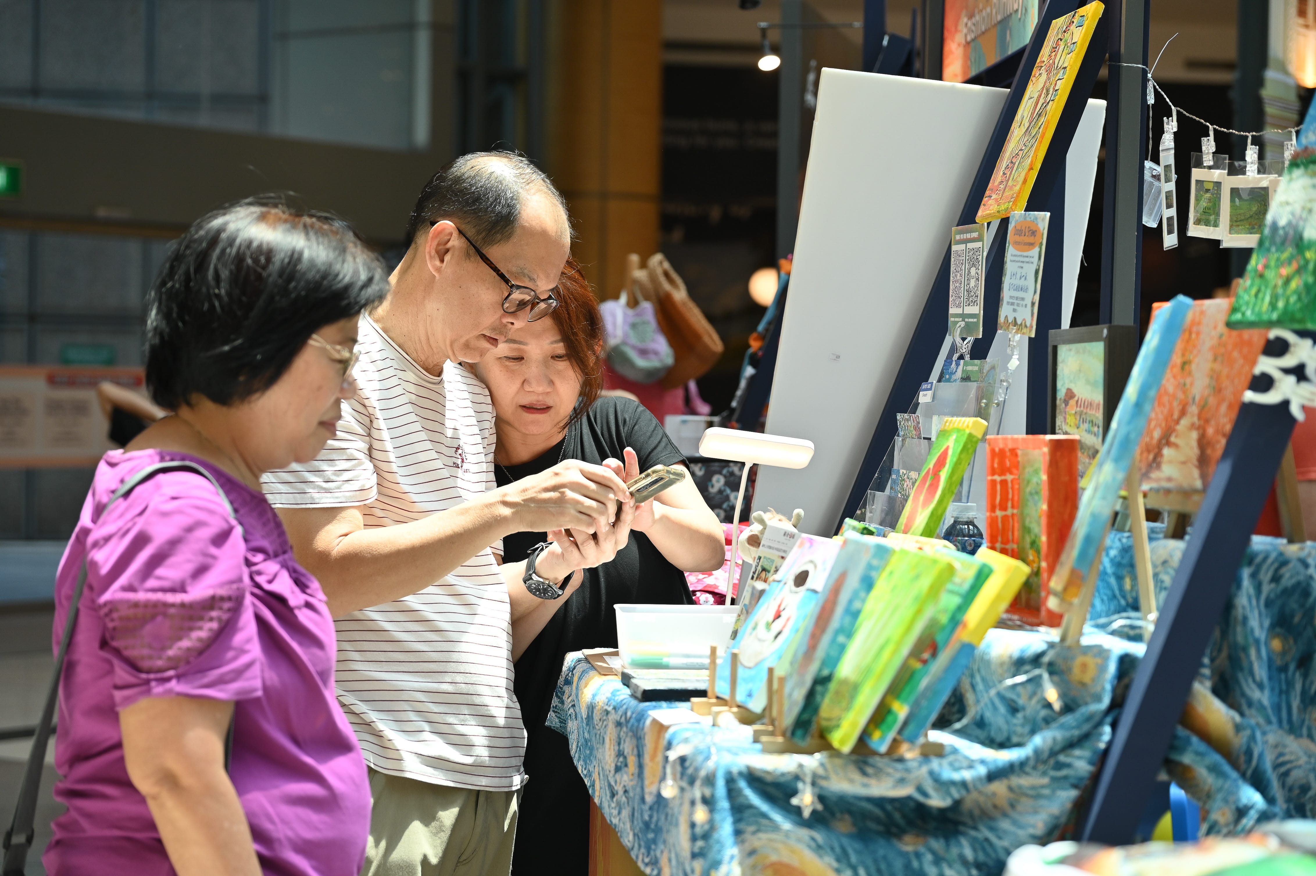 Two people are examining artworks displayed on a table in a brightly lit indoor space.