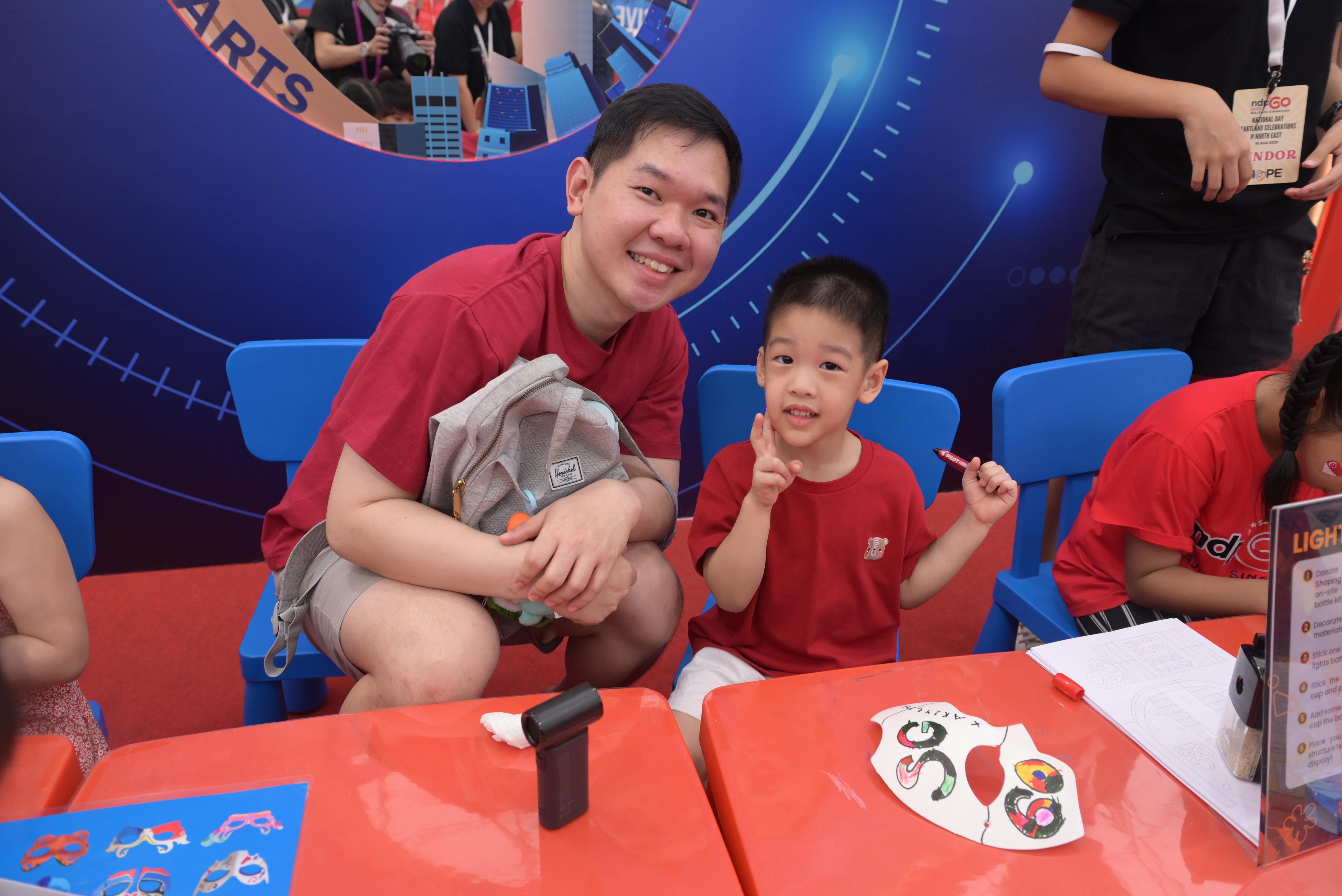 Two people in red shirts sit at brightly colored tables with blue chairs surrounded by art supplies and crafts.
