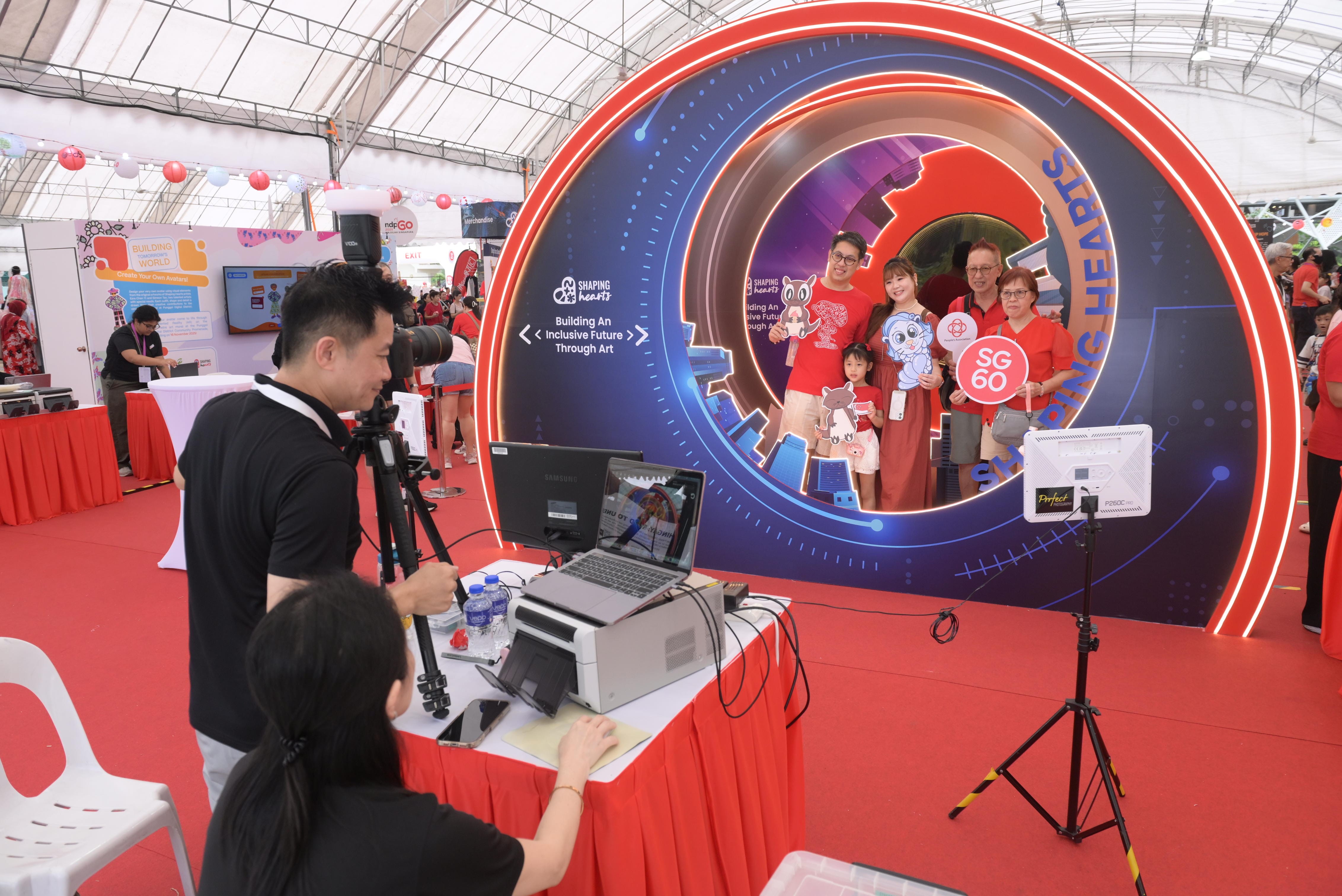 Two individuals operate a camera setup at the photobooth zone while a group stands inside a circular display with colourful decorations and signs.