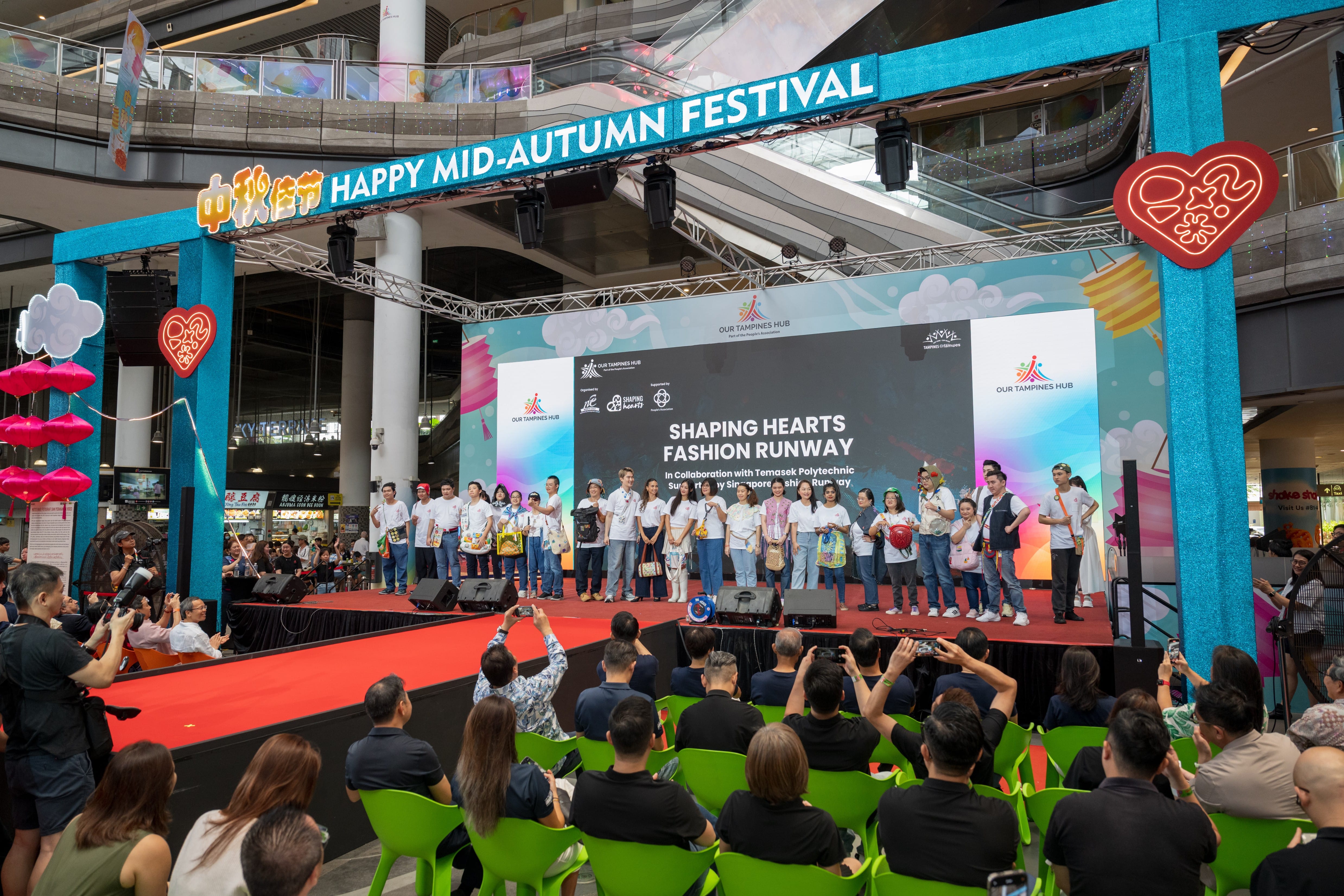 A group of people stand on stage under a "Happy Mid-Autumn Festival" sign at a fashion runway event with an audience front.