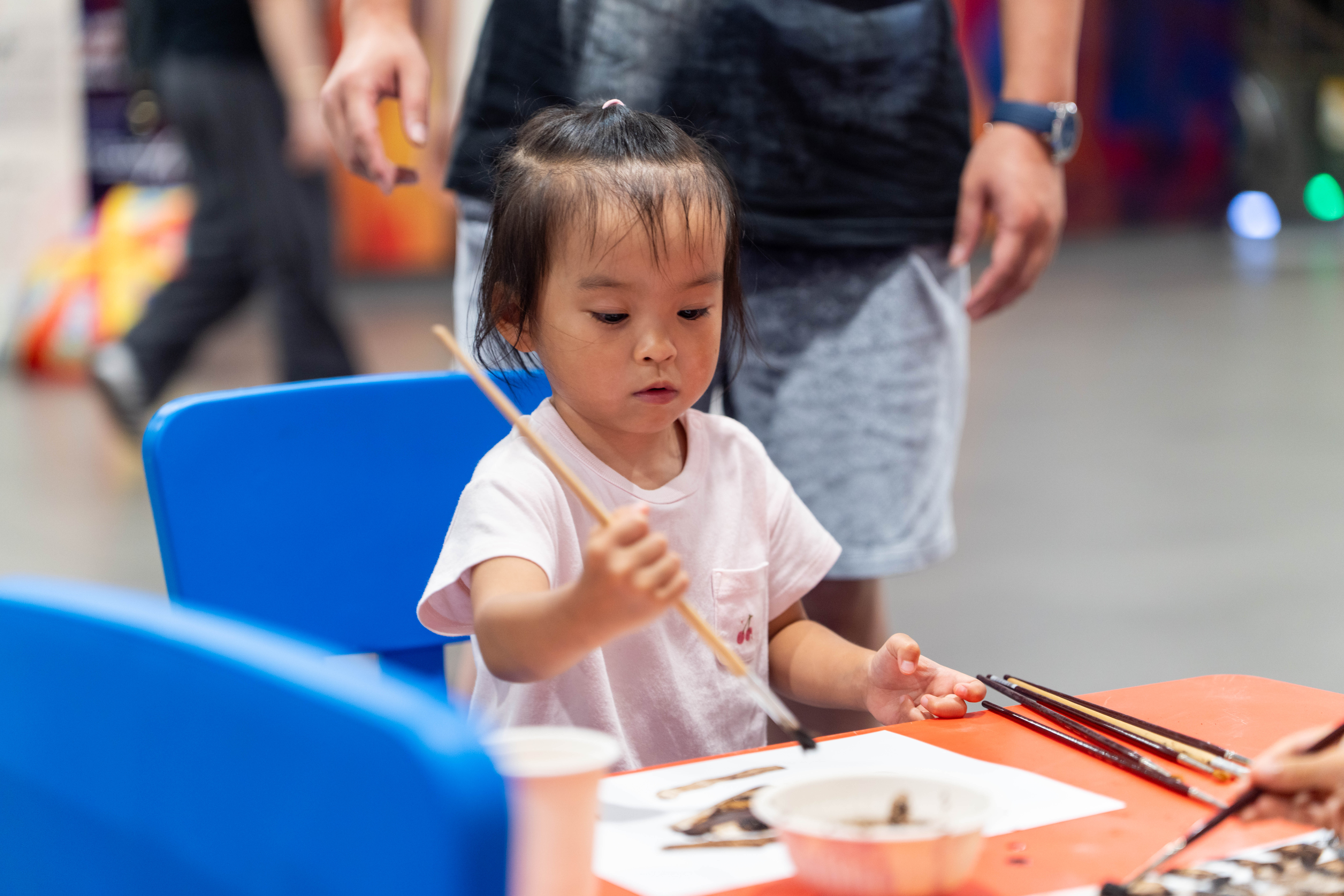 A child holding a paintbrush sits at a table with art supplies, including brushes and paper, while an adult stands nearby.