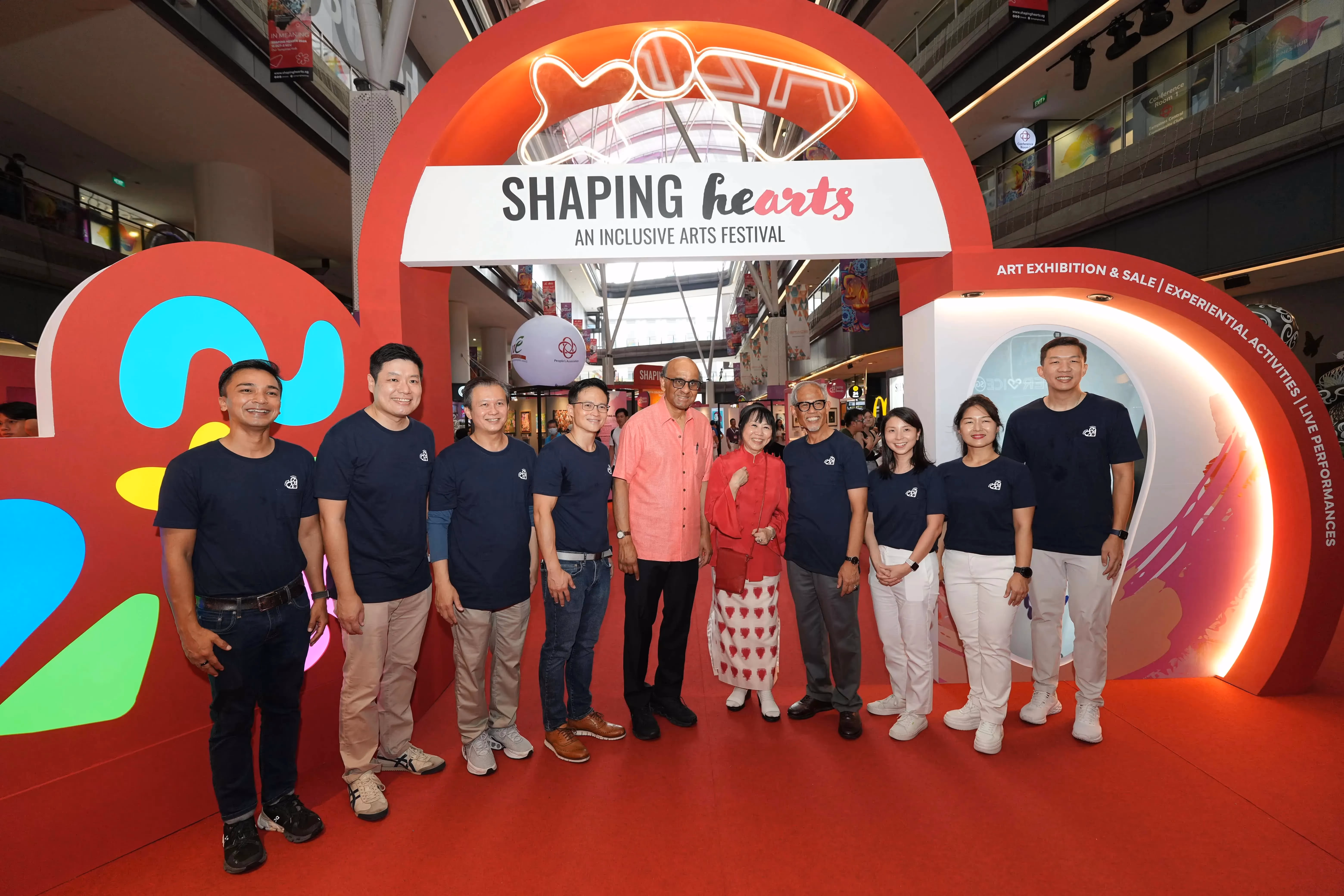 Ten people stand under a banner reading "Shaping Hearts: An Inclusive Arts Festival" in a brightly lit indoor space.