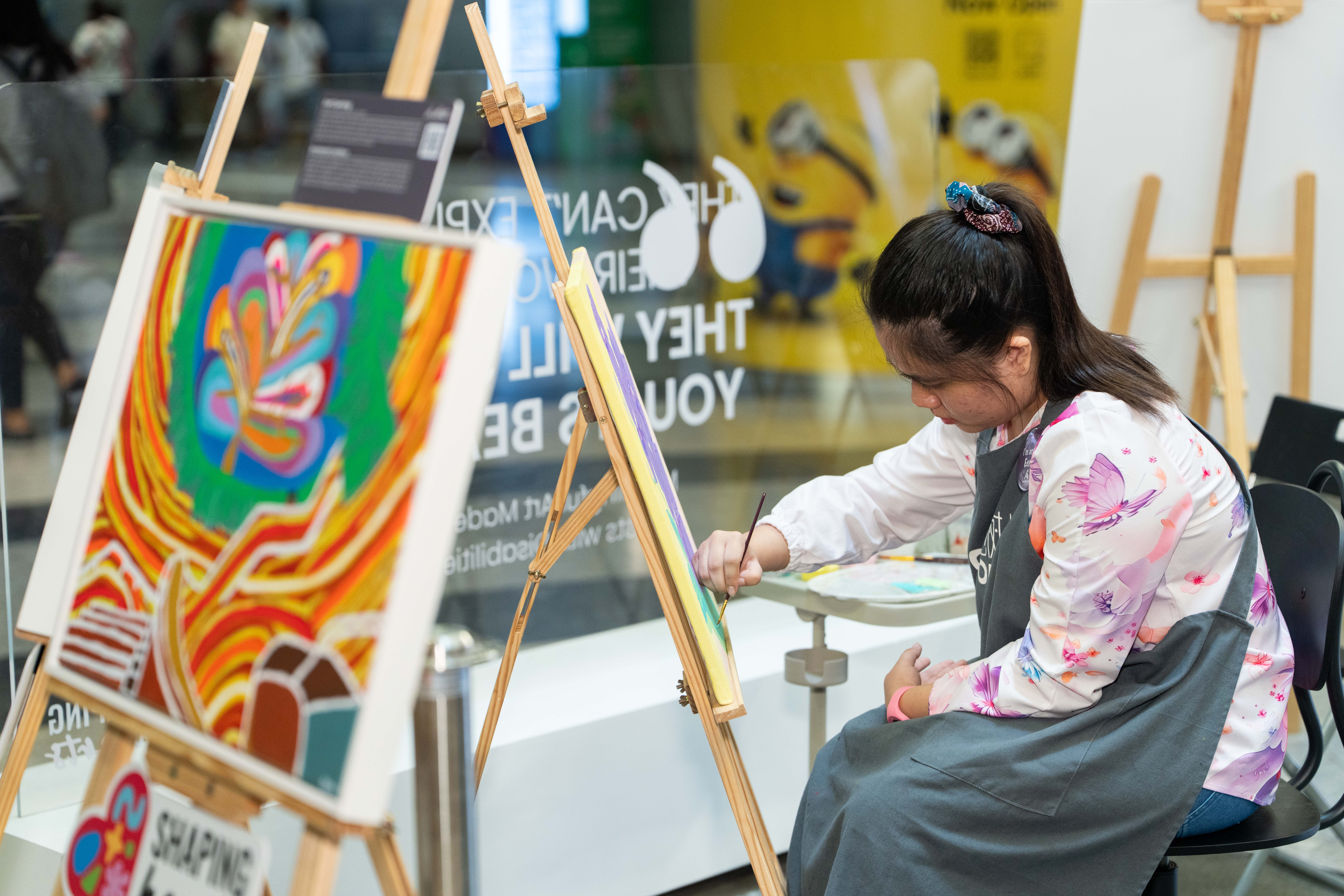 A person seated and painting on a canvas, with colourful artwork displayed on an easel nearby.