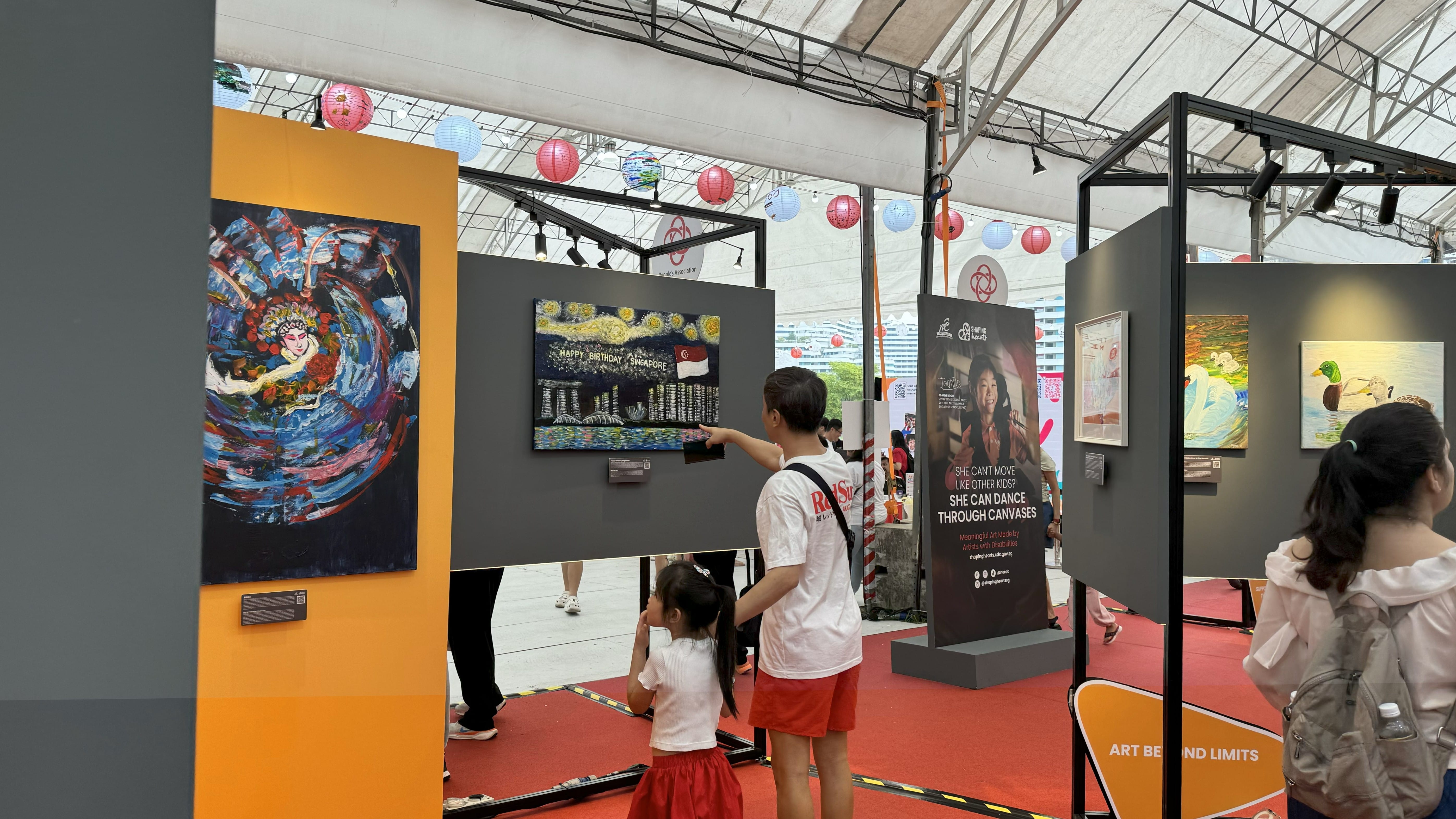 A father points at colourful paintings with his daughter in an art exhibit with lanterns hanging from the ceiling.