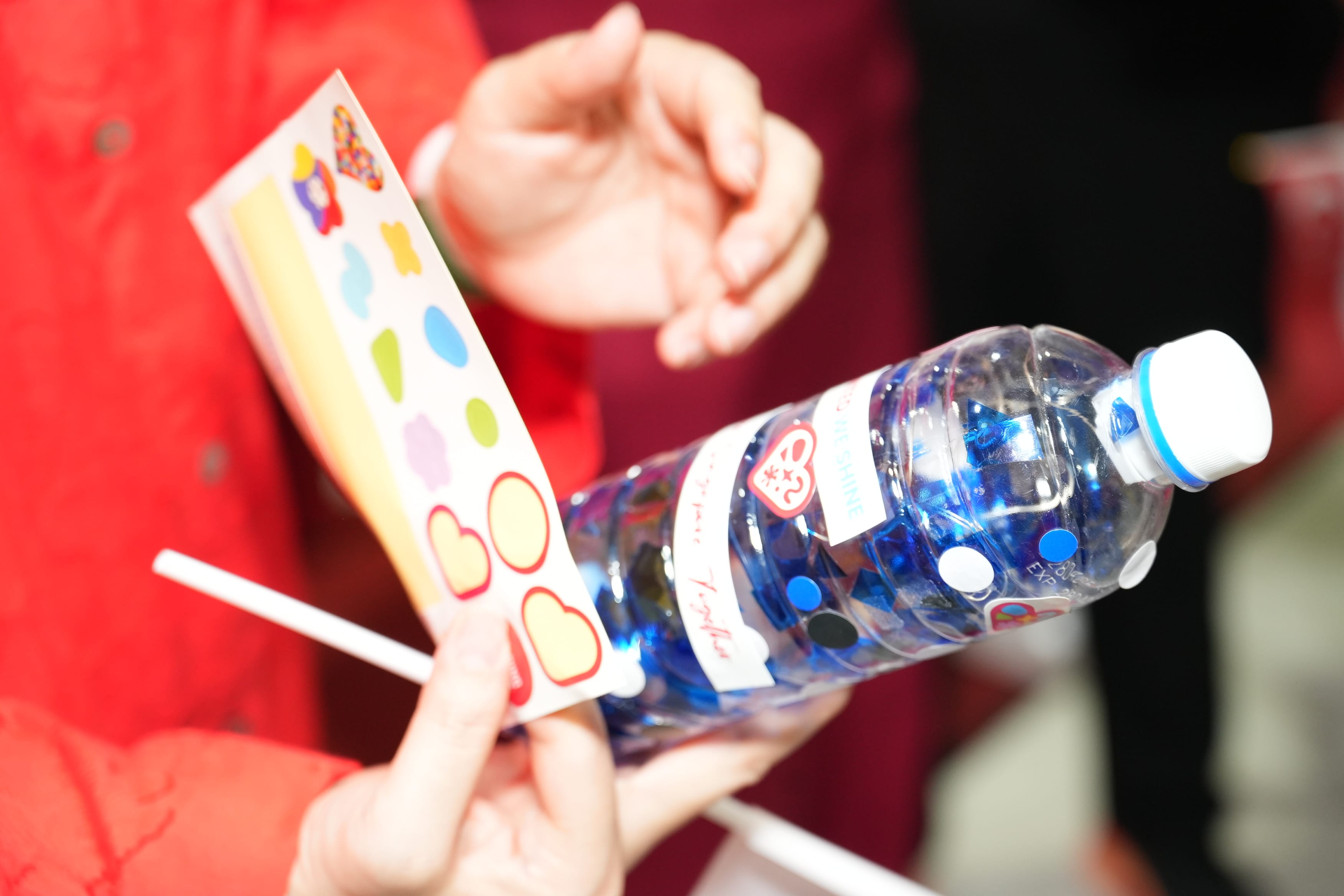 Hand holds a water bottle with blue confetti inside, alongside a sheet of colorful stickers.
