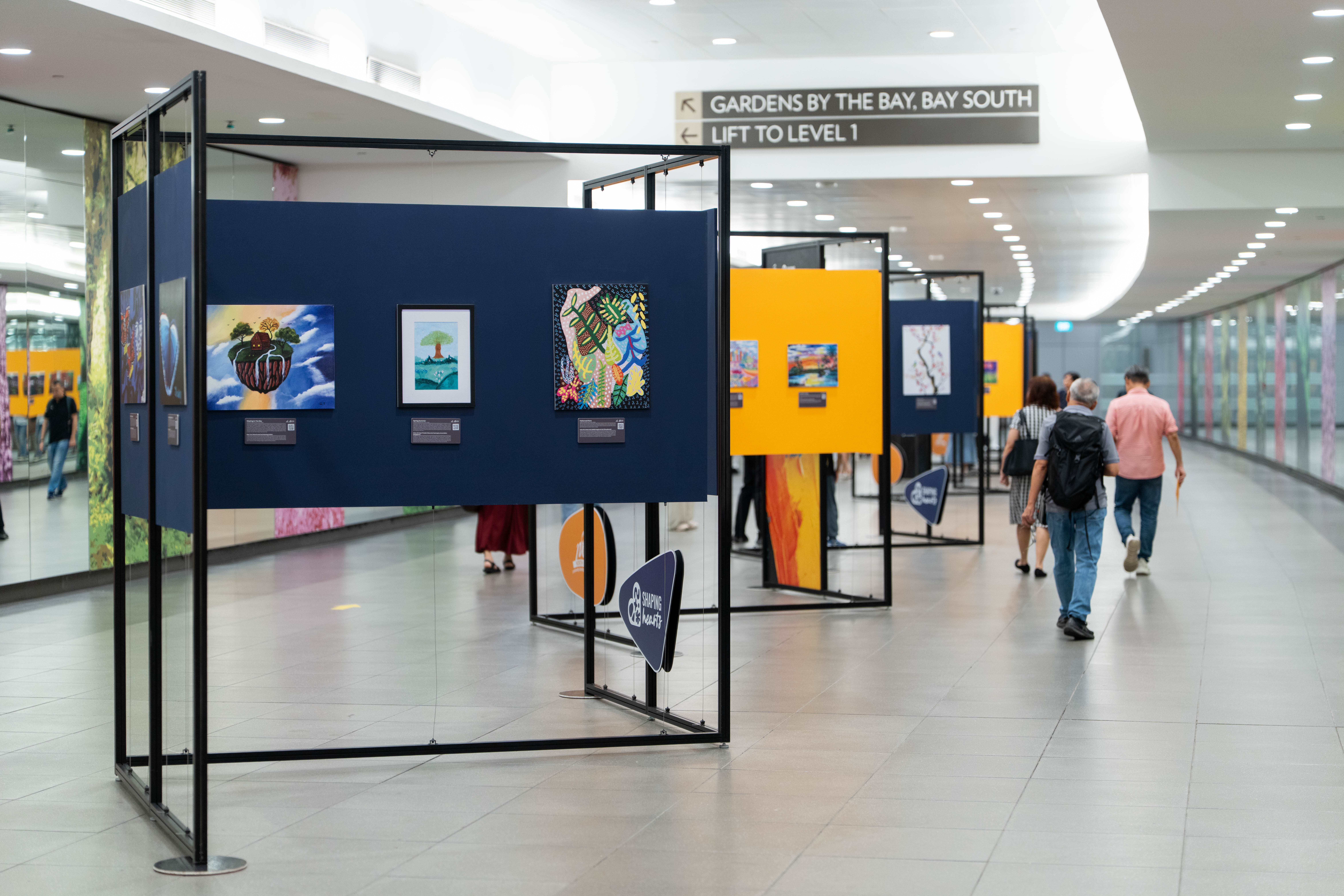 People walk past art displays in a brightly lit hallway, with paintings on blue and yellow panels.