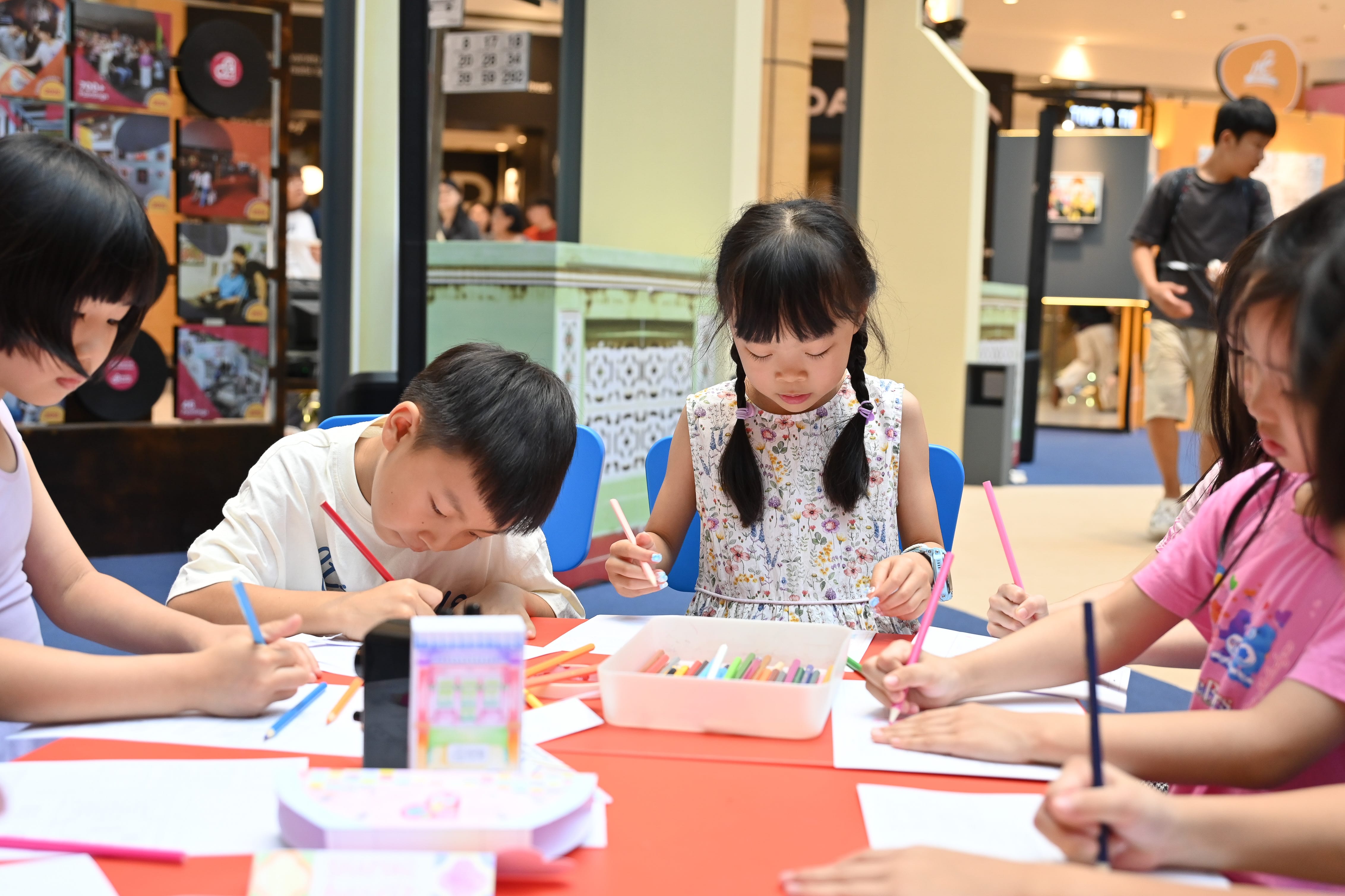 Children sitting at a table colouring with crayons and markers in an indoor setting with other people in the background.
