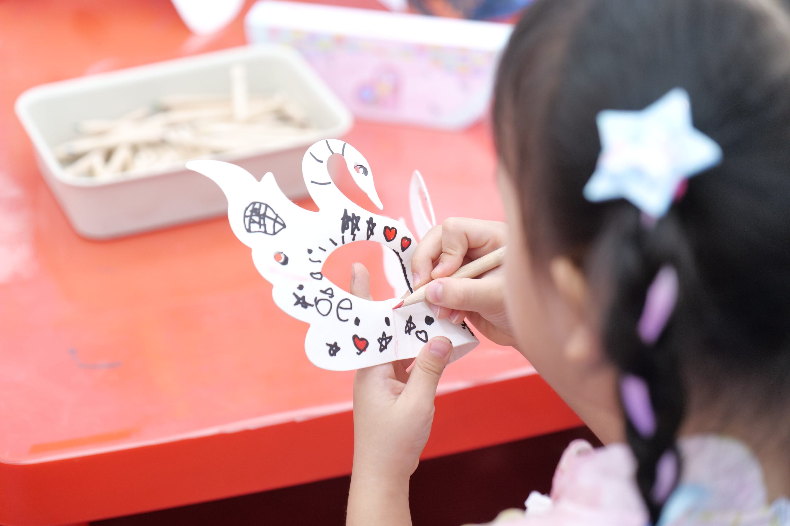 A child with braided hair decorates a white paper mask with black and red drawings at a red table.