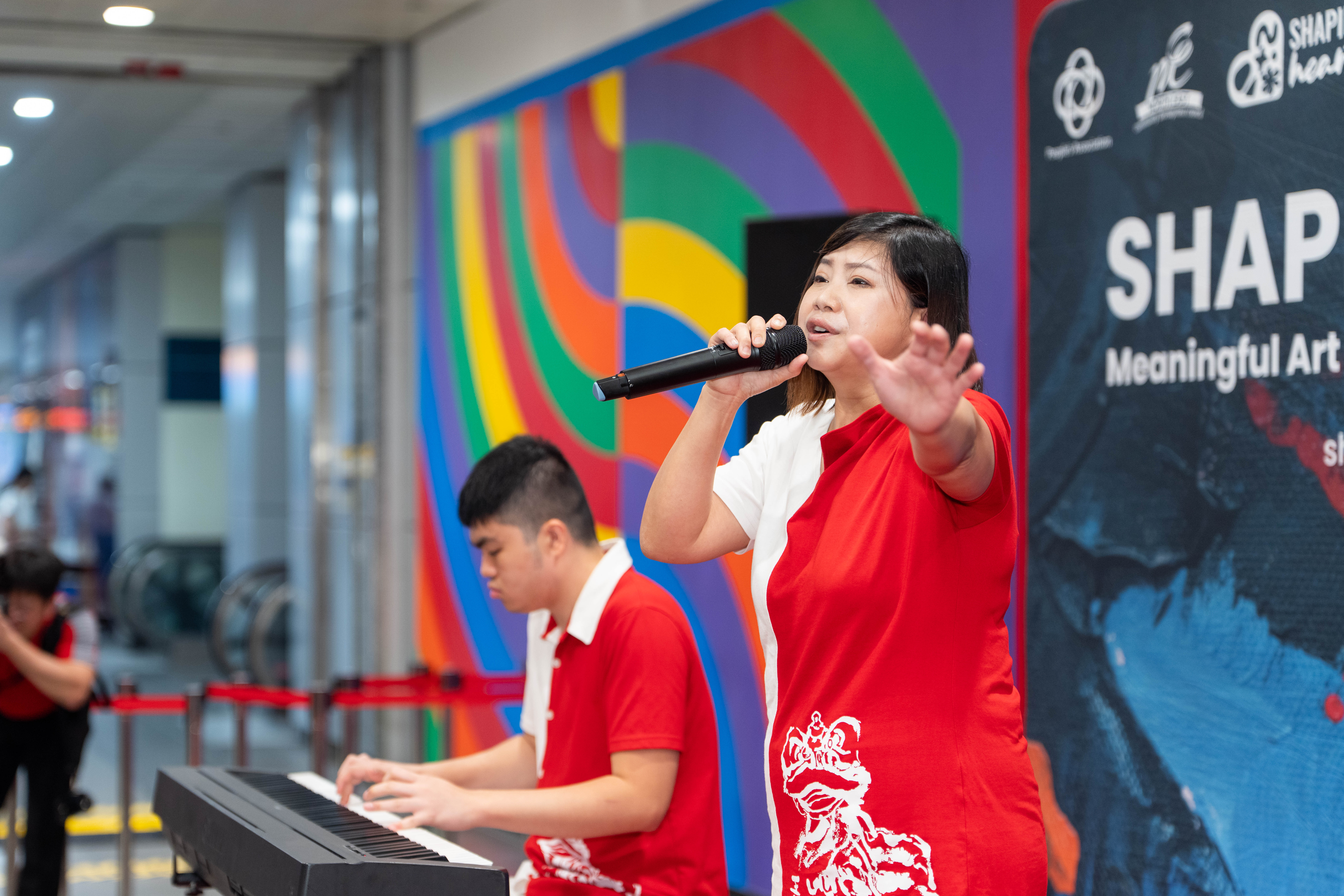 A person in red and white clothes sings into a microphone while another plays keyboard in front of a colorful wall.