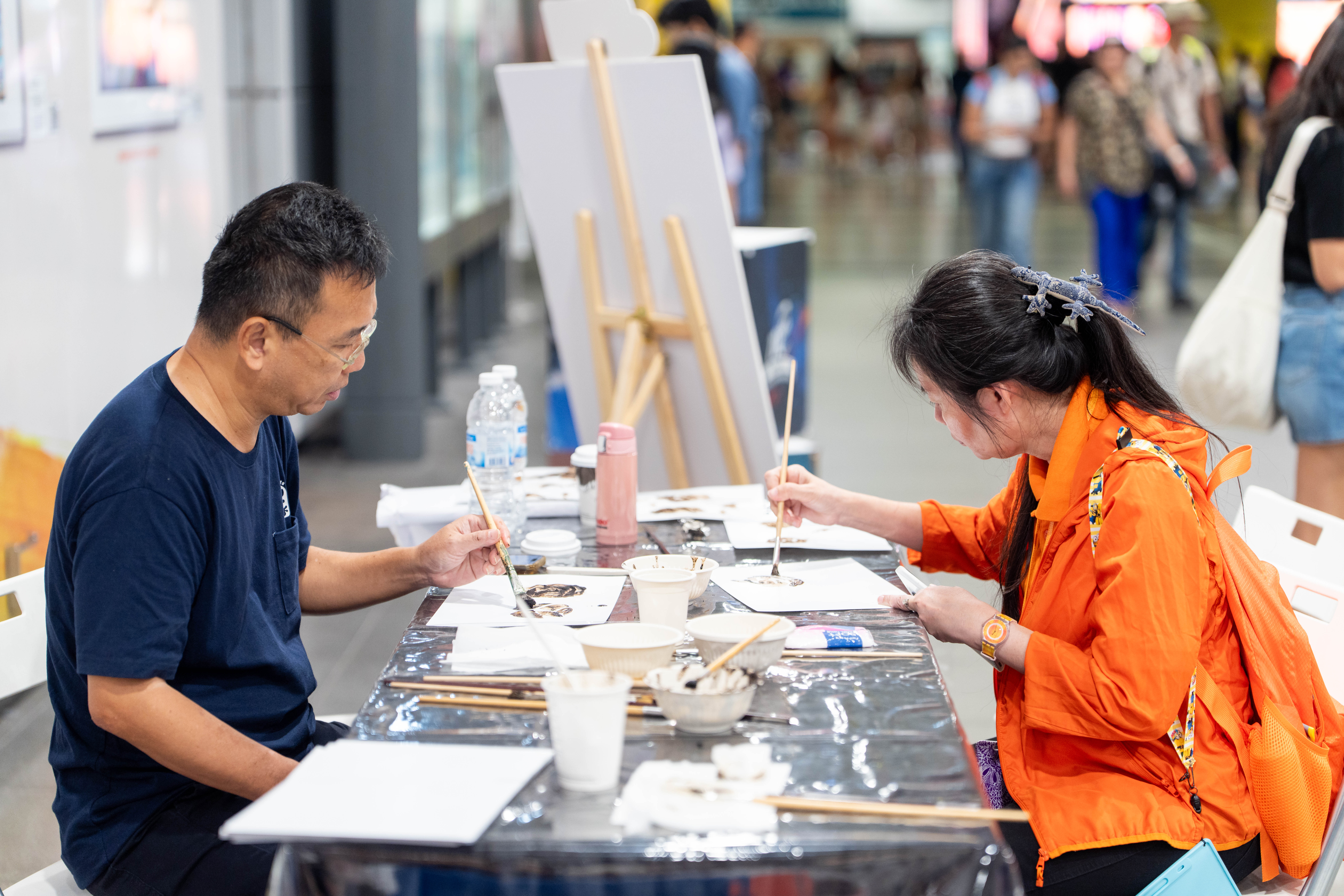 Two people sit at a table painting with brushes, surrounded by art supplies and a wooden easel.