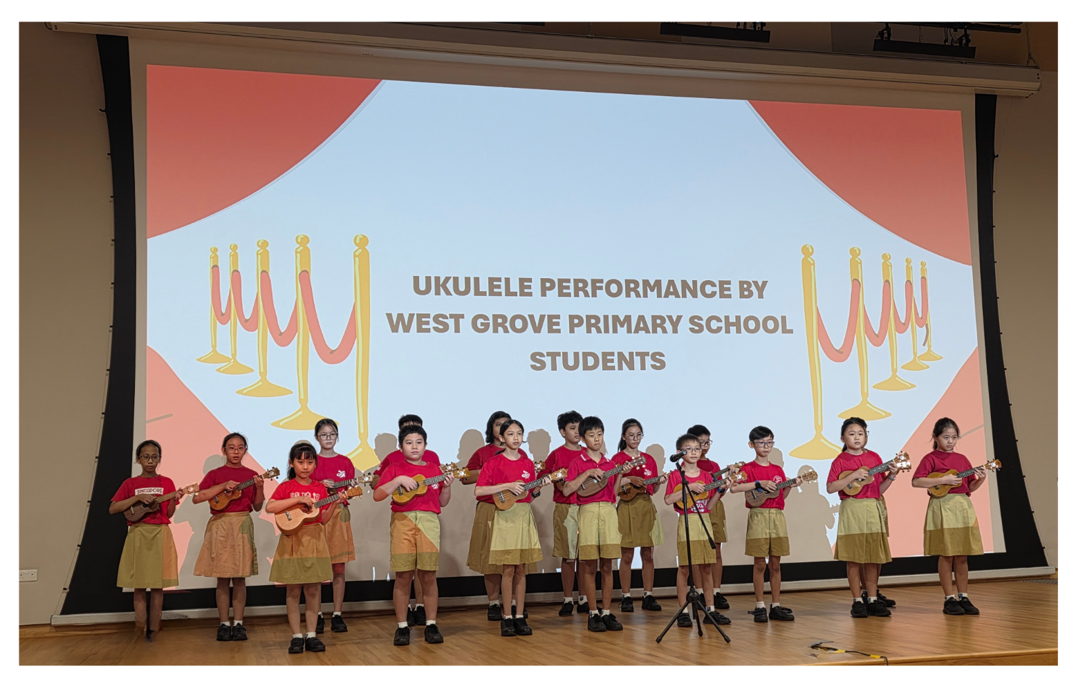 Children wearing red shirts and yellow shorts stand on stage playing ukuleles; screen text is "Ukulele Performance by West Grove Primary School Students".