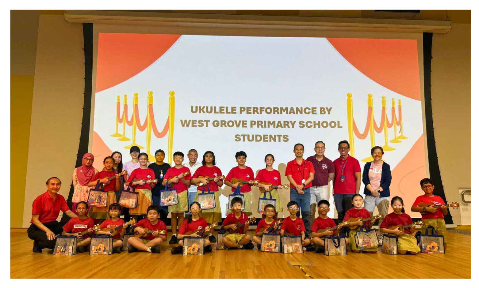 Students and adults in red shirts pose with ukuleles in front of a projected sign about a performance at West Grove Primary School.