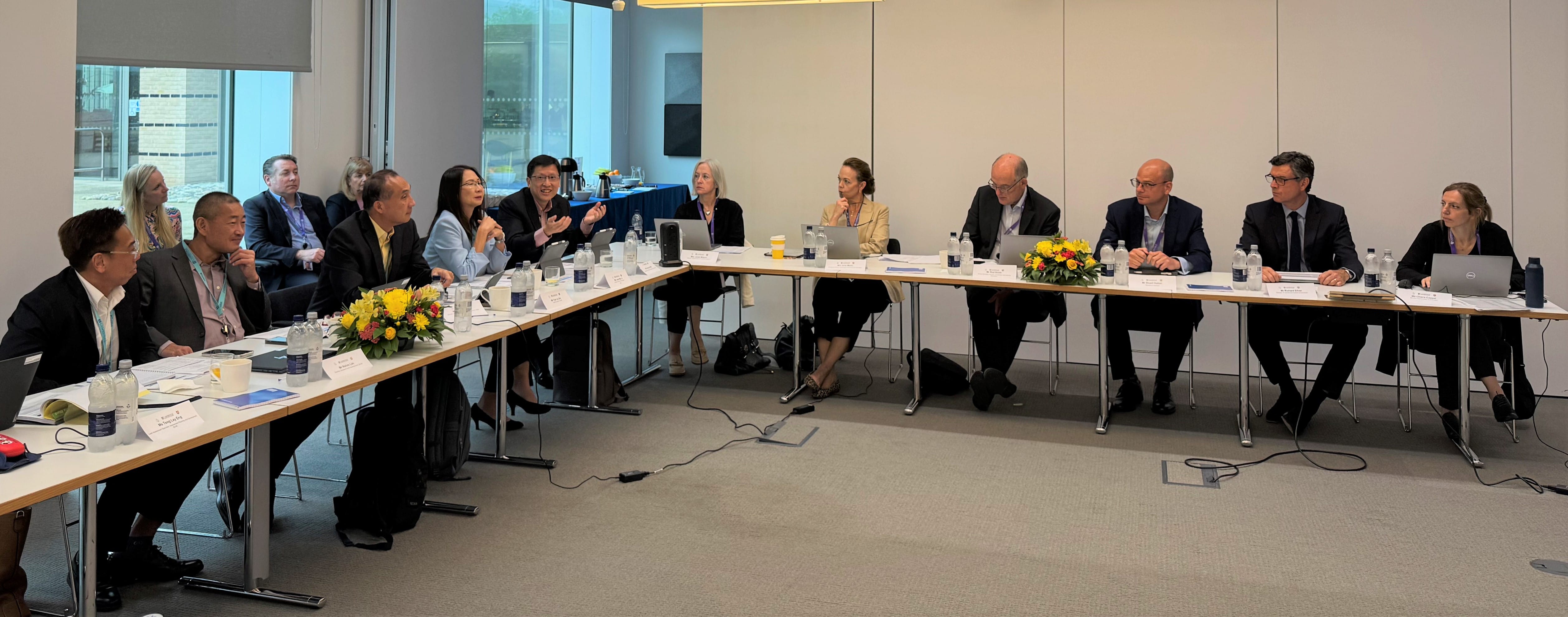 People are seated around a large rectangular table, engaging in discussions, with laptops, documents, and flower arrangements present.