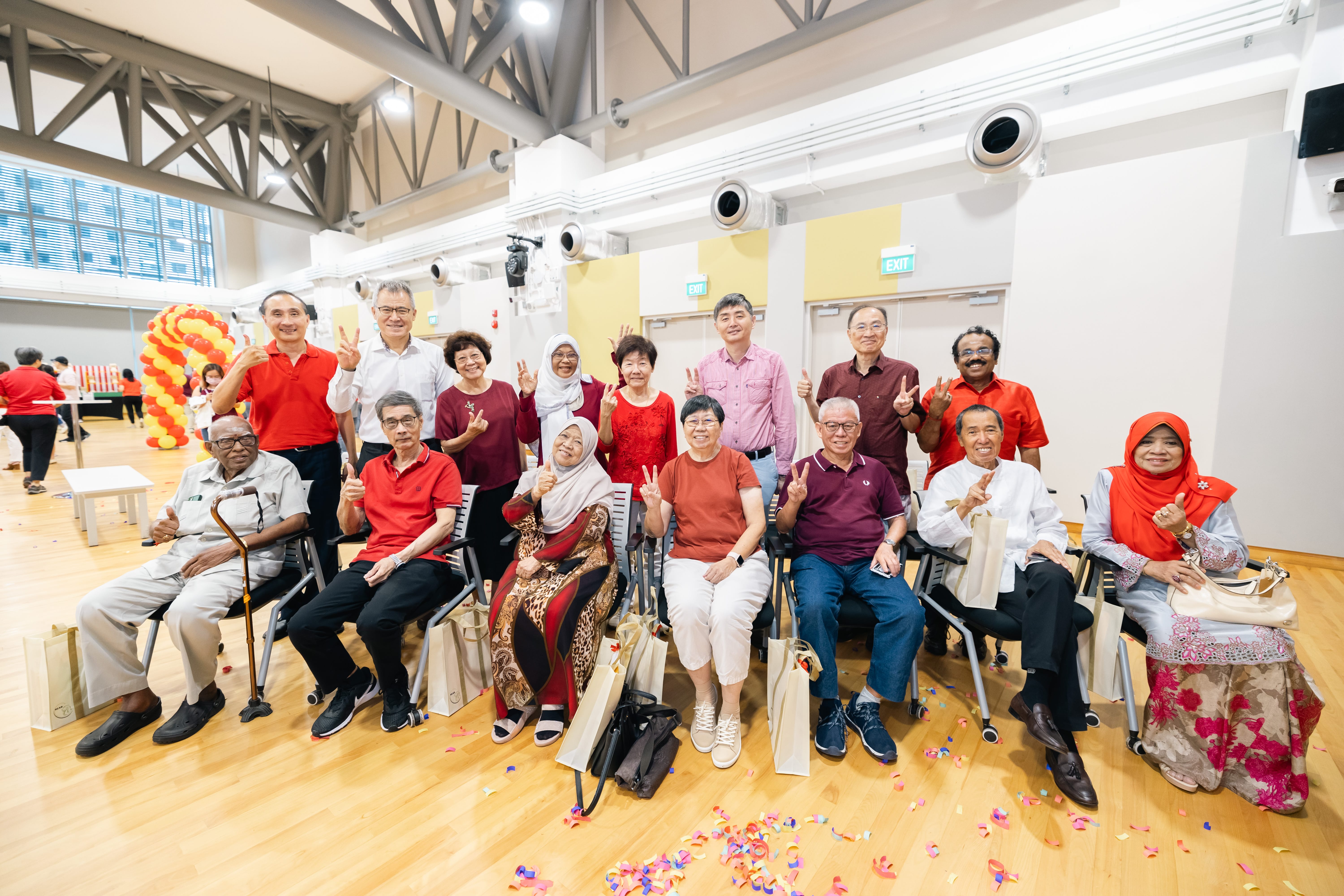 A group of people in colorful clothes pose indoors, some holding peace signs; balloons and confetti are visible in the background.
