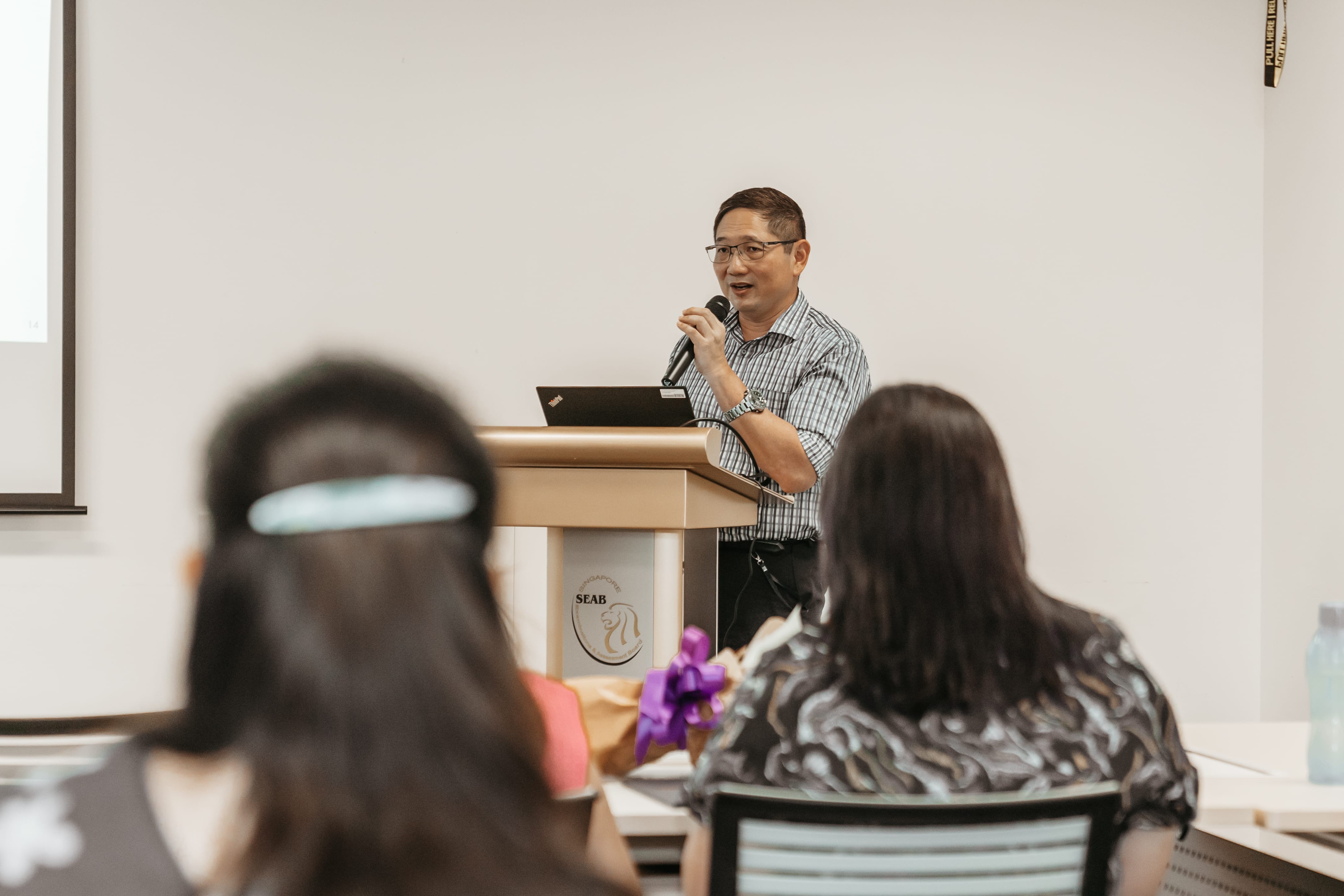 A person in a striped shirt is speaking at a podium with a laptop in front of an audience in a classroom setting.