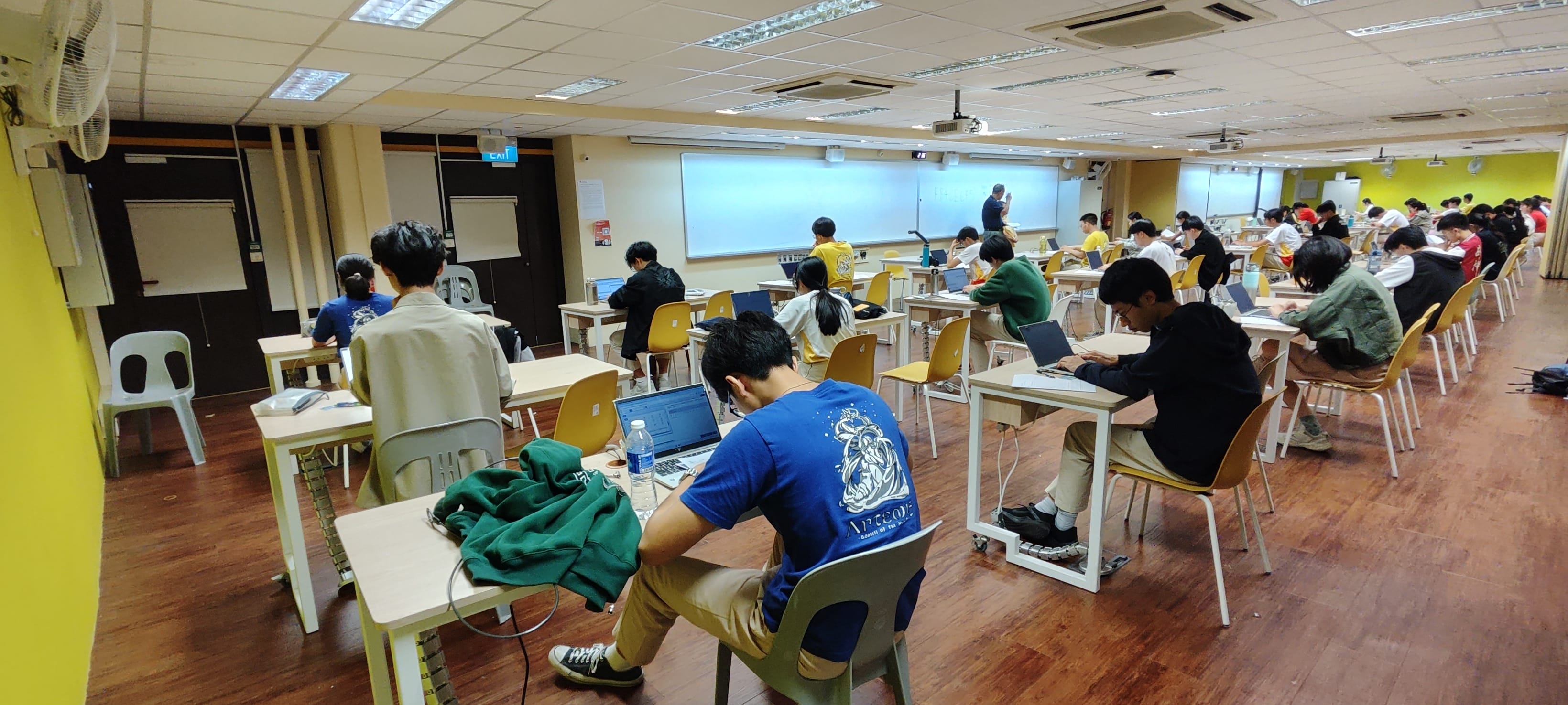 Classroom filled with students sitting at desks, using laptops, and facing a lecturer at the front.