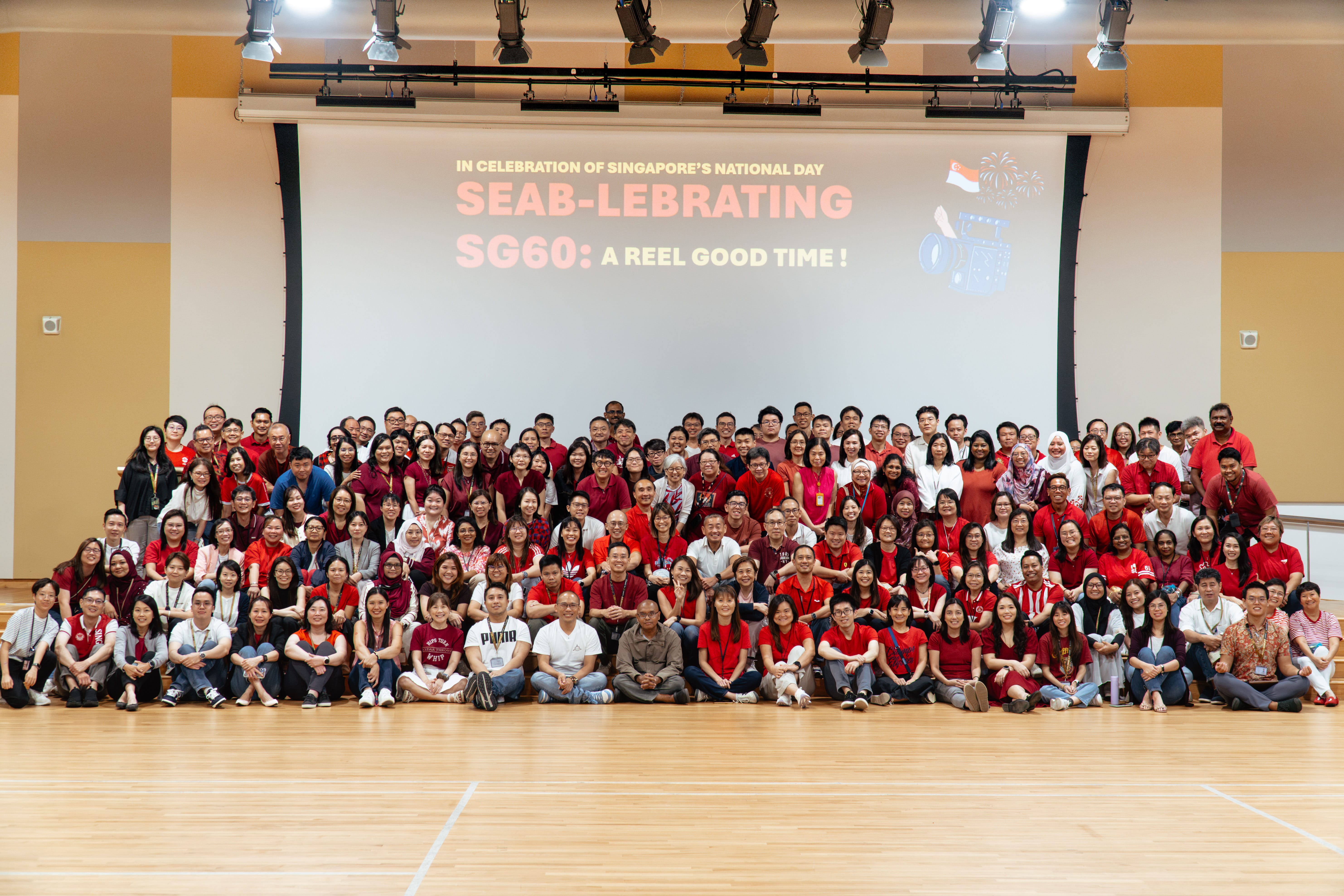 A large group of people in red and white attire posed in front of a screen with text celebrating Singapore's National Day.