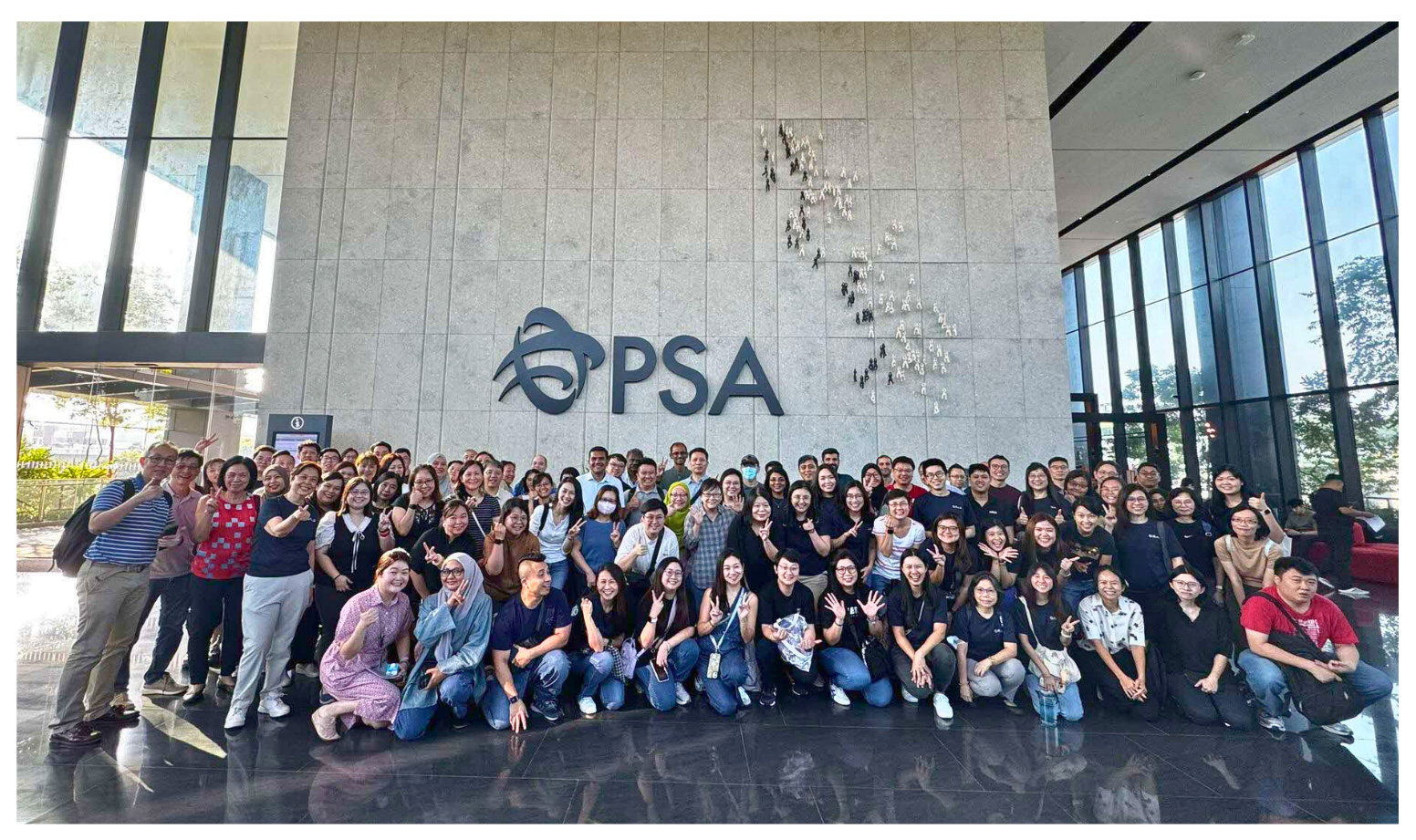A large group of people posing indoors in front of a sign that reads "PSA" on a grey wall.