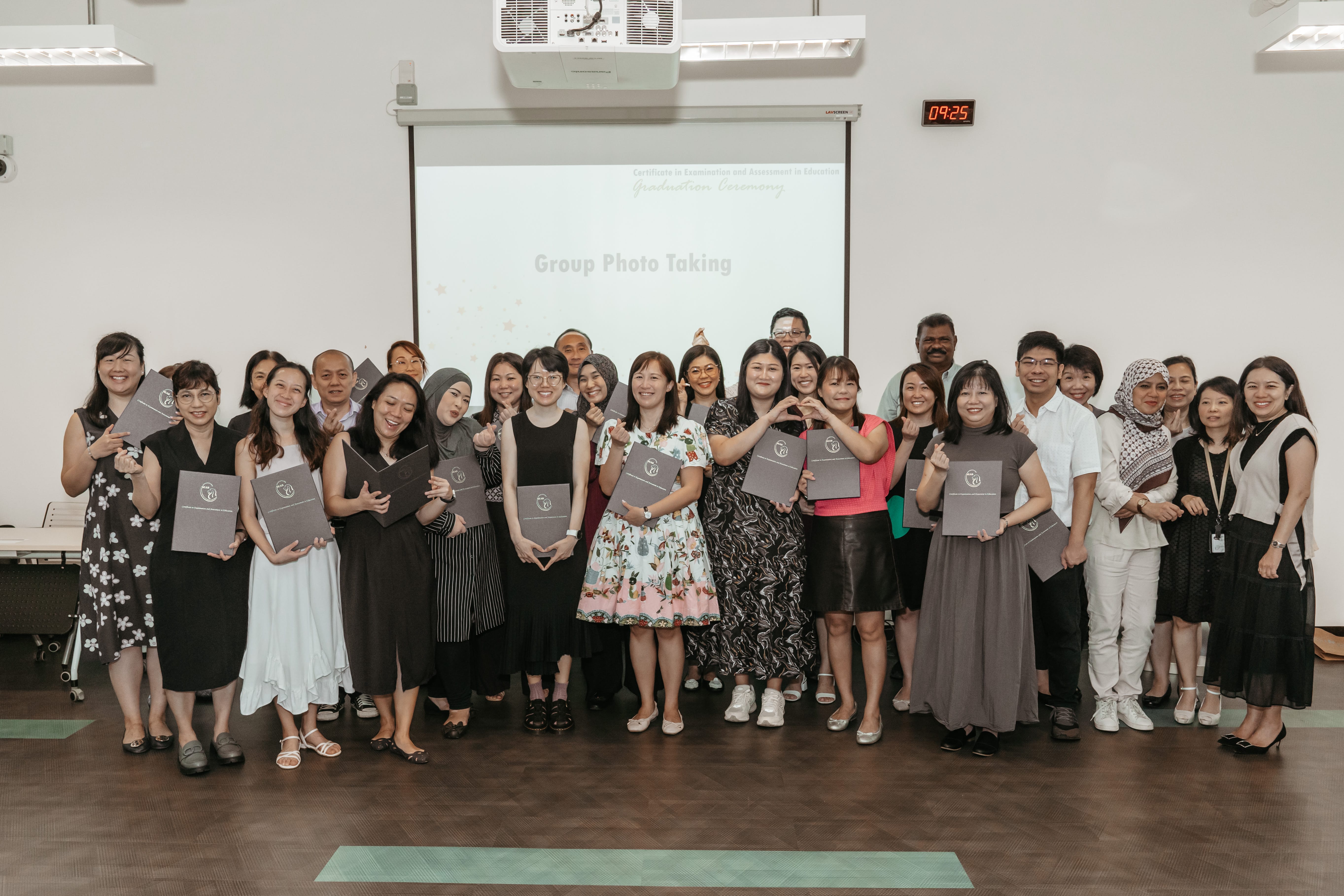 A group of people stand indoors holding certificates, with "Group Photo Taking" displayed on a screen behind them.