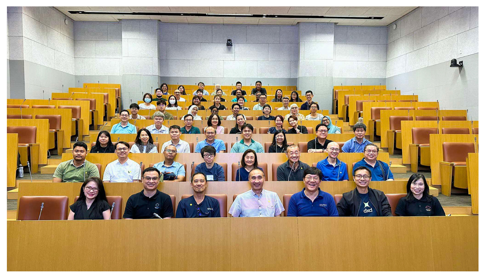 A large group of people are seated in a tiered lecture hall with wooden desks and microphones in front of them.