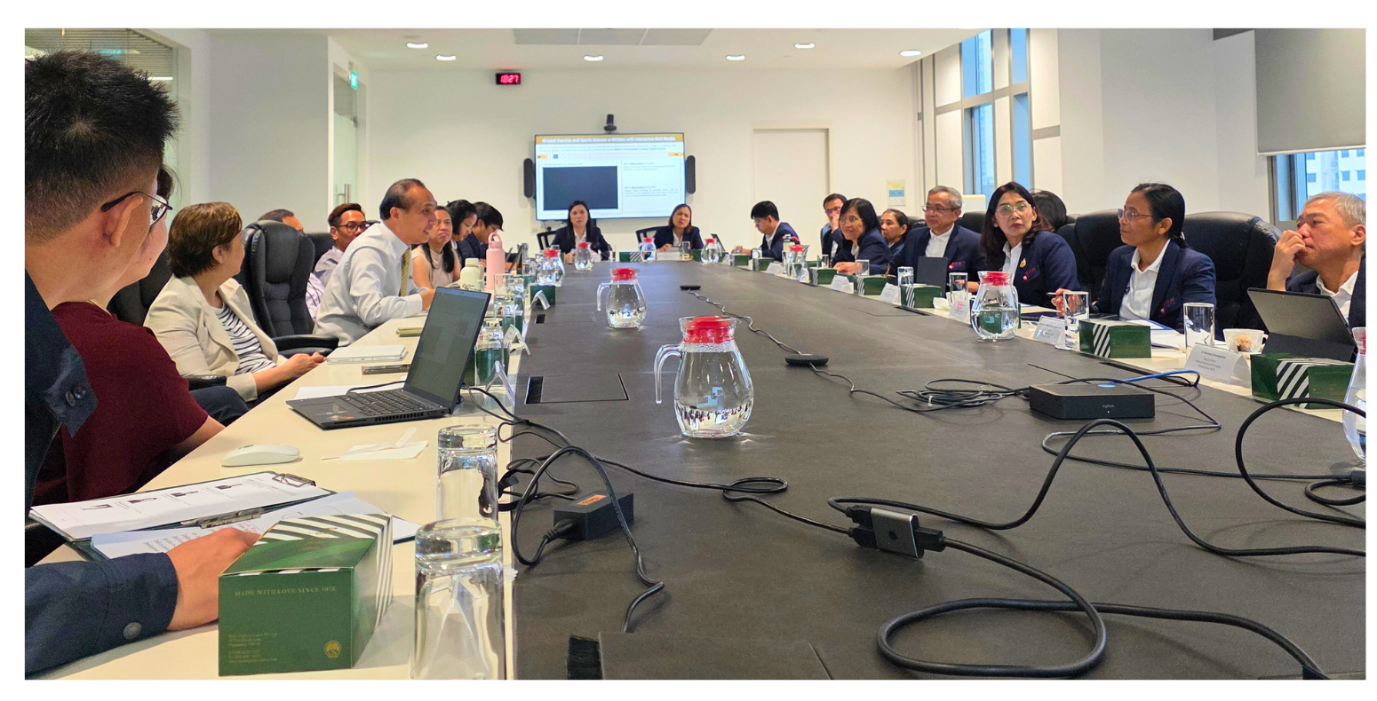 People are seated around a large conference table with laptops and documents, facing a screen displaying a presentation.