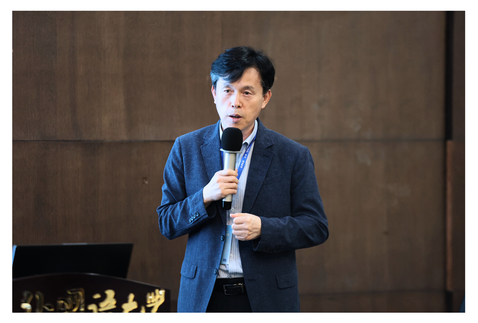 A person in a dark blazer holds a microphone in front of a wooden wall.