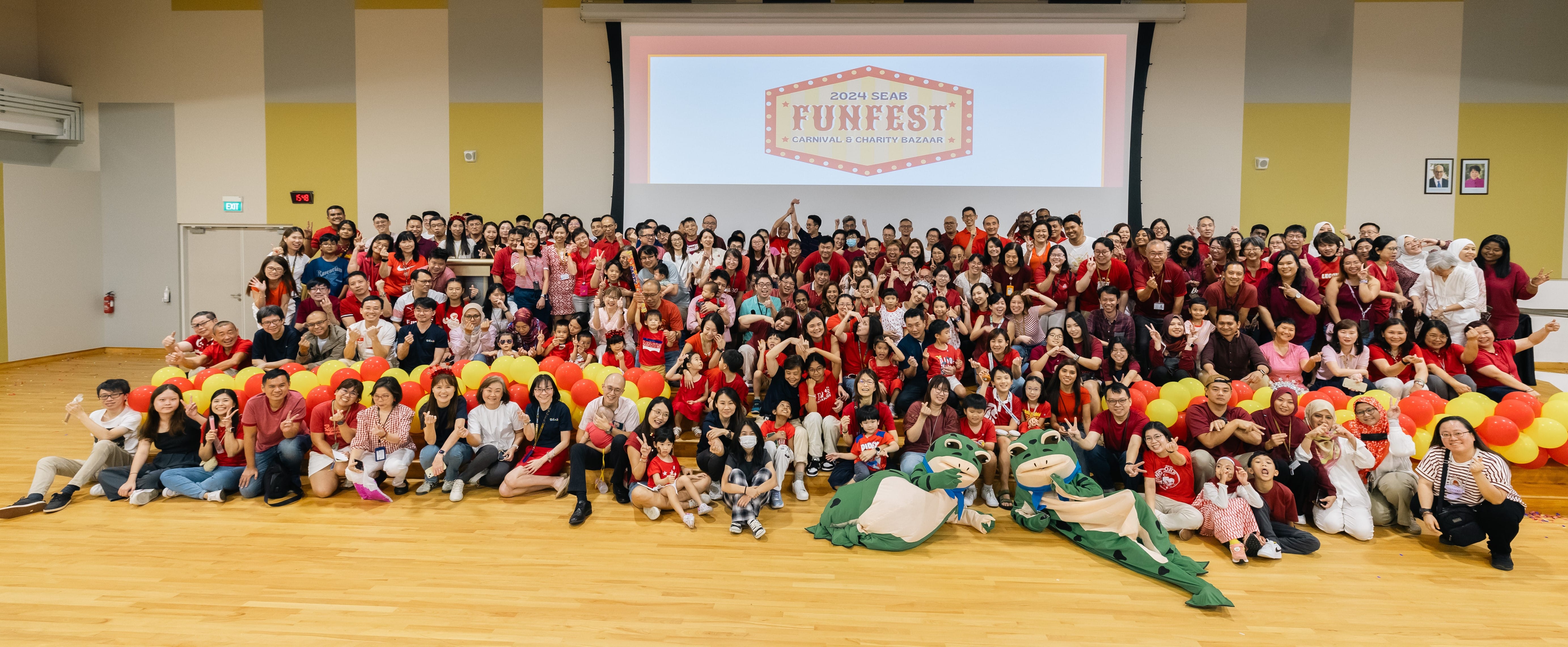 Large group of people posing indoors near red and yellow balloons, with "2024 SEAB Funfest: Carnival & Charity Bazaar" on a screen.