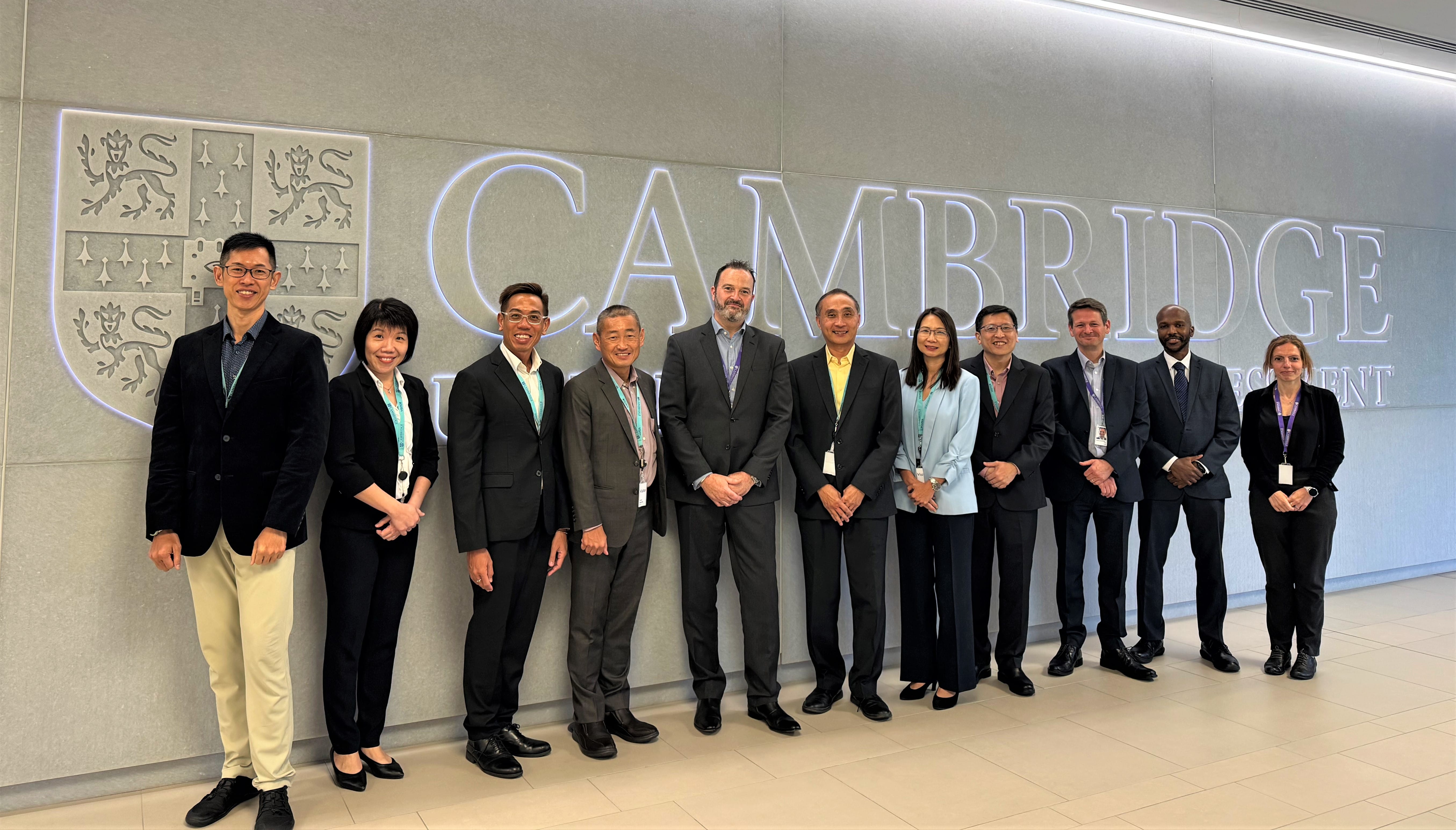 A group of people in formal attire stands in front of a wall with the word "CAMBRIDGE" and a shield emblem.