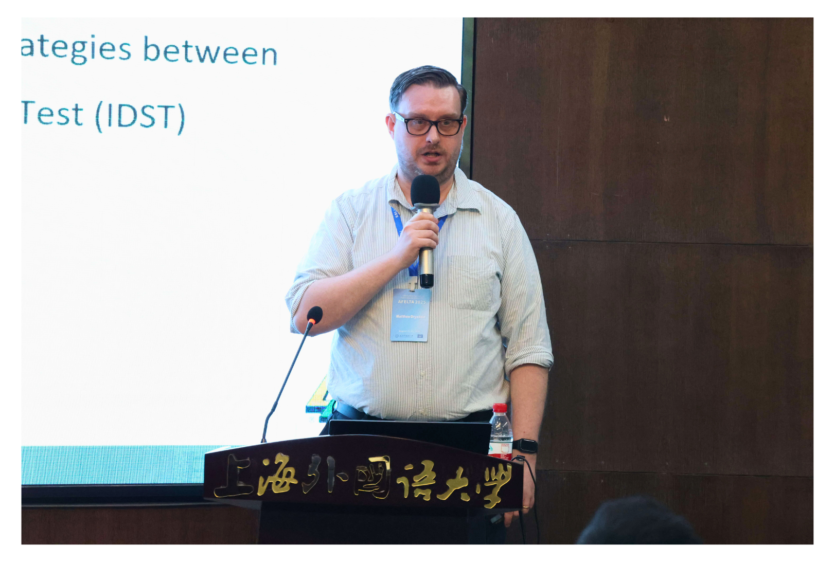 A person in a blue shirt stands at a podium with a microphone, giving a presentation; part of the screen reads "Test (IDST)".
