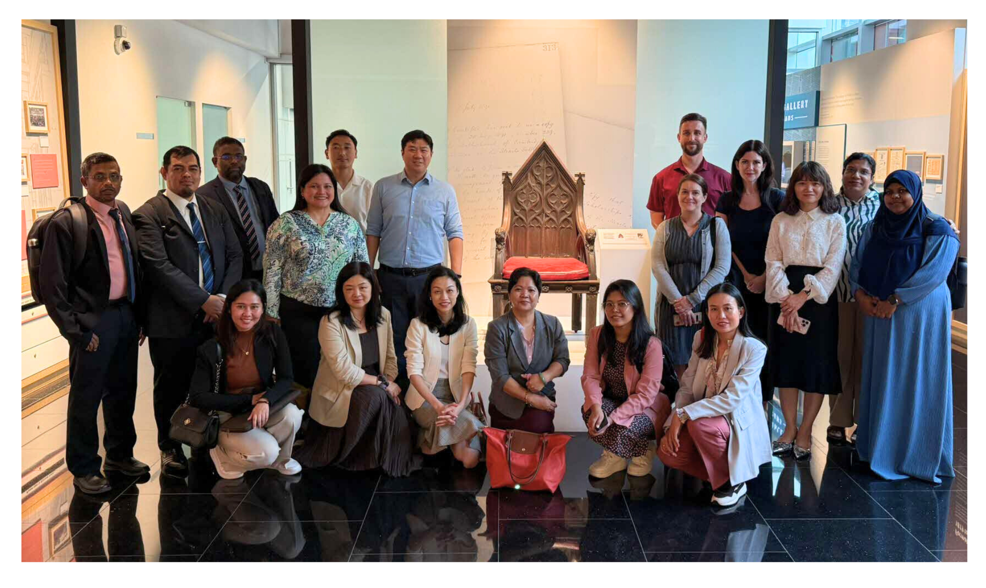A group of people are posing in front of an ornate chair with a red cushion, in a modern gallery setting.