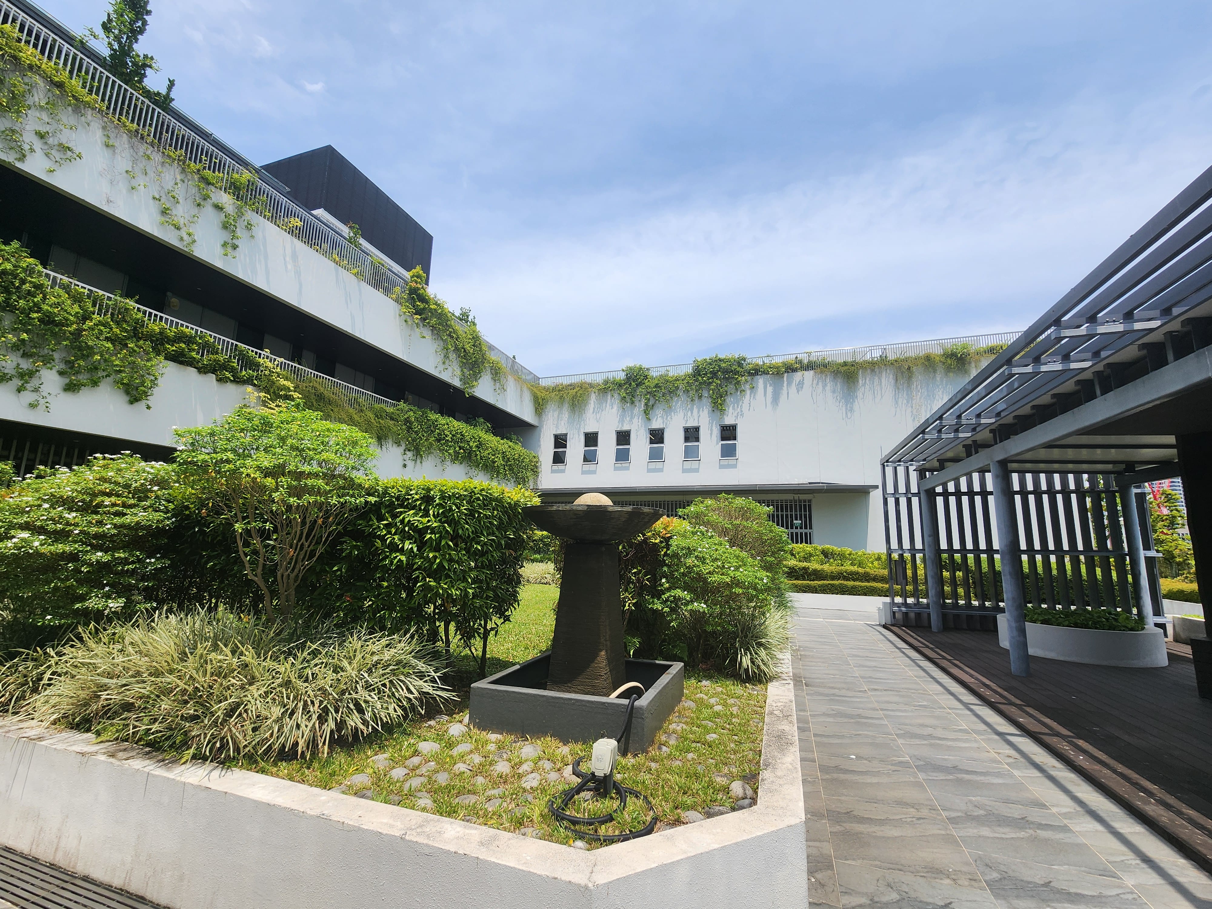 A courtyard with greenery, a central stone structure, and adjacent buildings with plants covering parts of their walls.