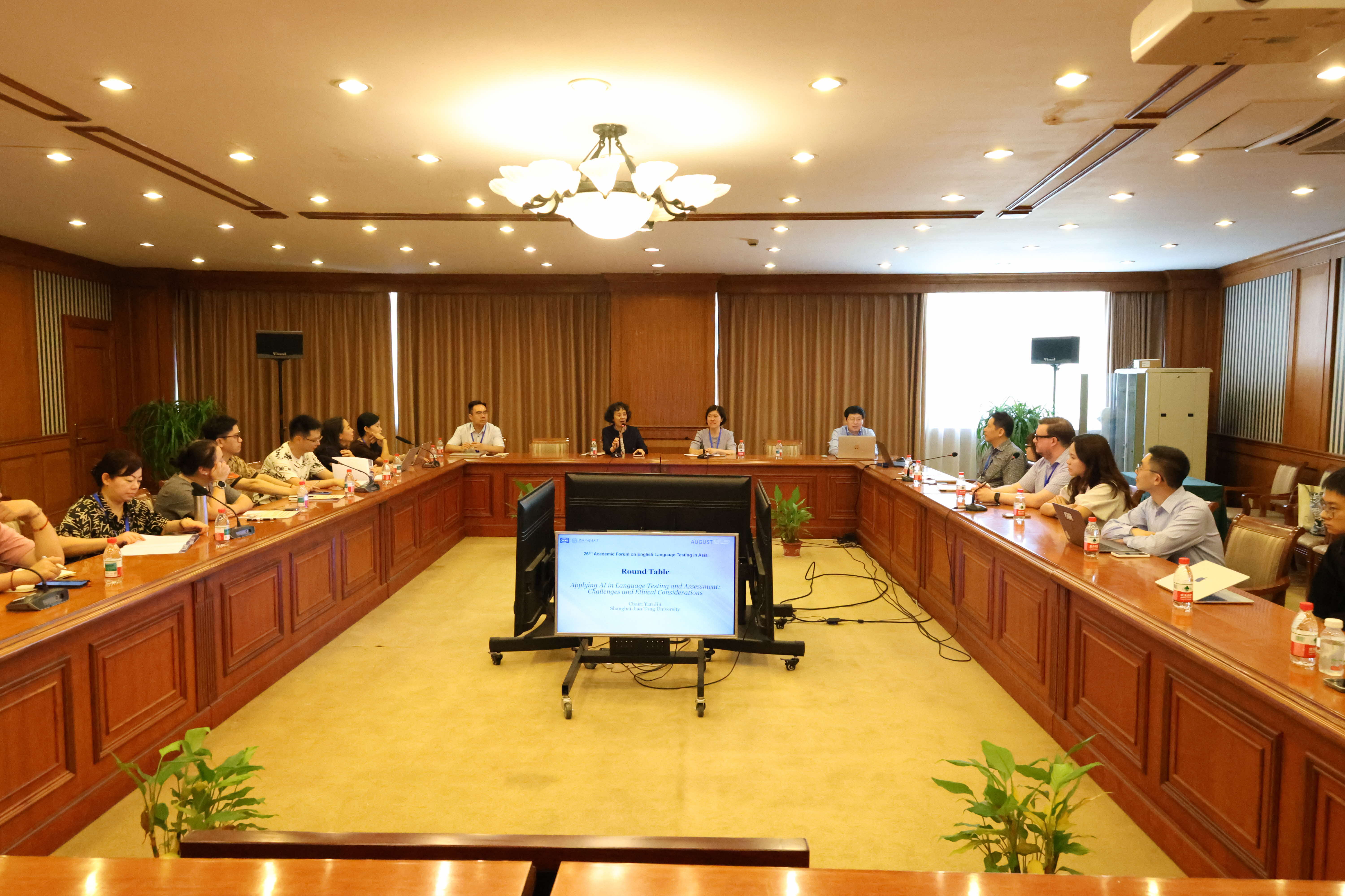 A conference room with people seated around a large U-shaped table, with two screens displaying text and plants in the foreground.