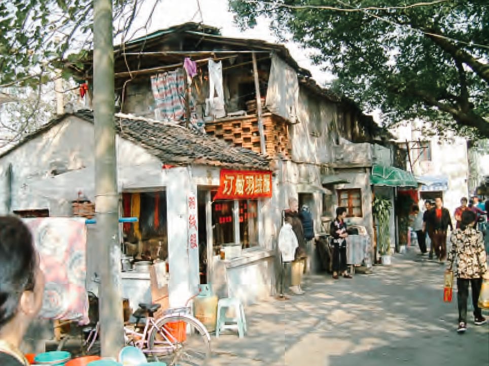 A narrow street lined with old Chinese shophouses, red signs, and people walking under large trees.