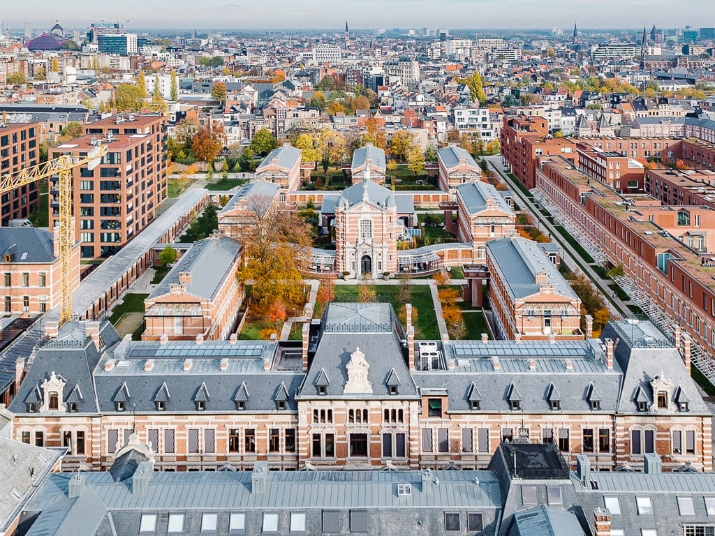 Aerial view of a large historic red brick complex with courtyards, autumn trees, and a dense European city surrounding it.