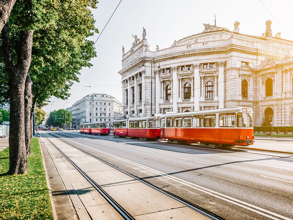 Red trams travel along a wide boulevard beside a grand white neoclassical building with columns and statues.