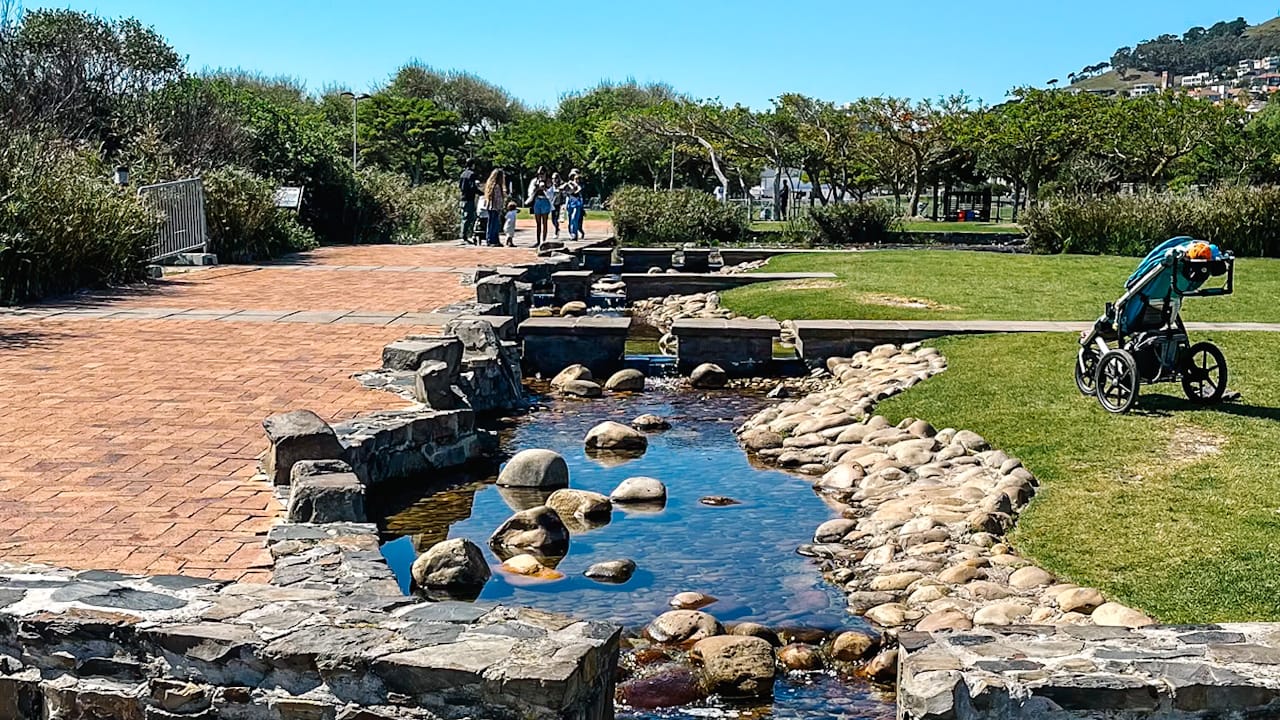 A rocky stream runs through a sunny park with a brick path, green lawn, stroller, and people walking among trees.