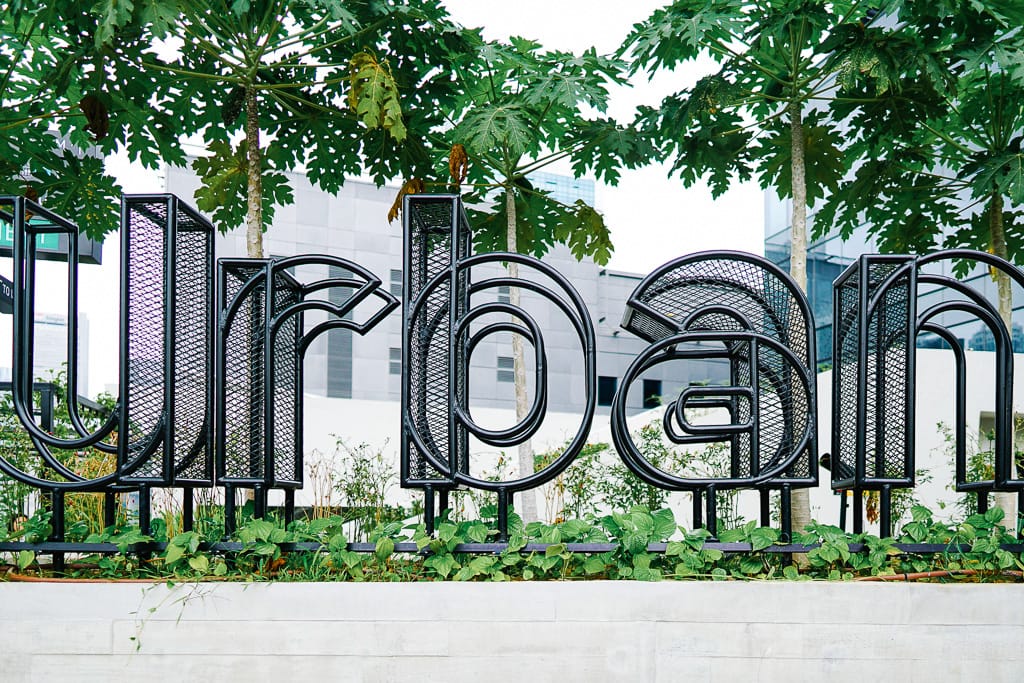 Large black metal letters spelling "urban" stand in a garden bed with green plants and tall trees behind them.
