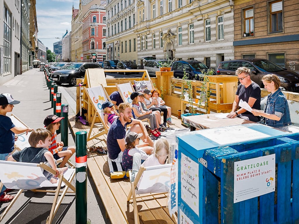 Adults and children relax on wooden deck chairs in a temporary street parklet labeled Gratzloase in a city.
